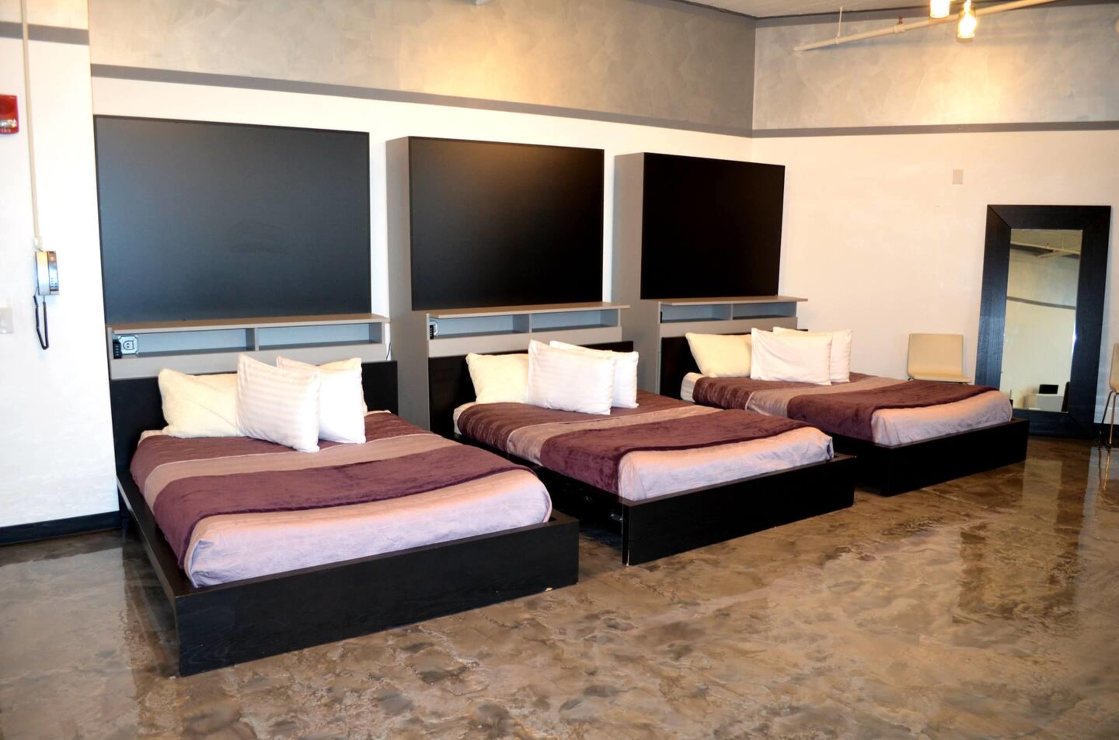 A simple room containing three twin beds with dark wood frames, each made with light bedding and positioned parallel to a wall with two large black panels above them.