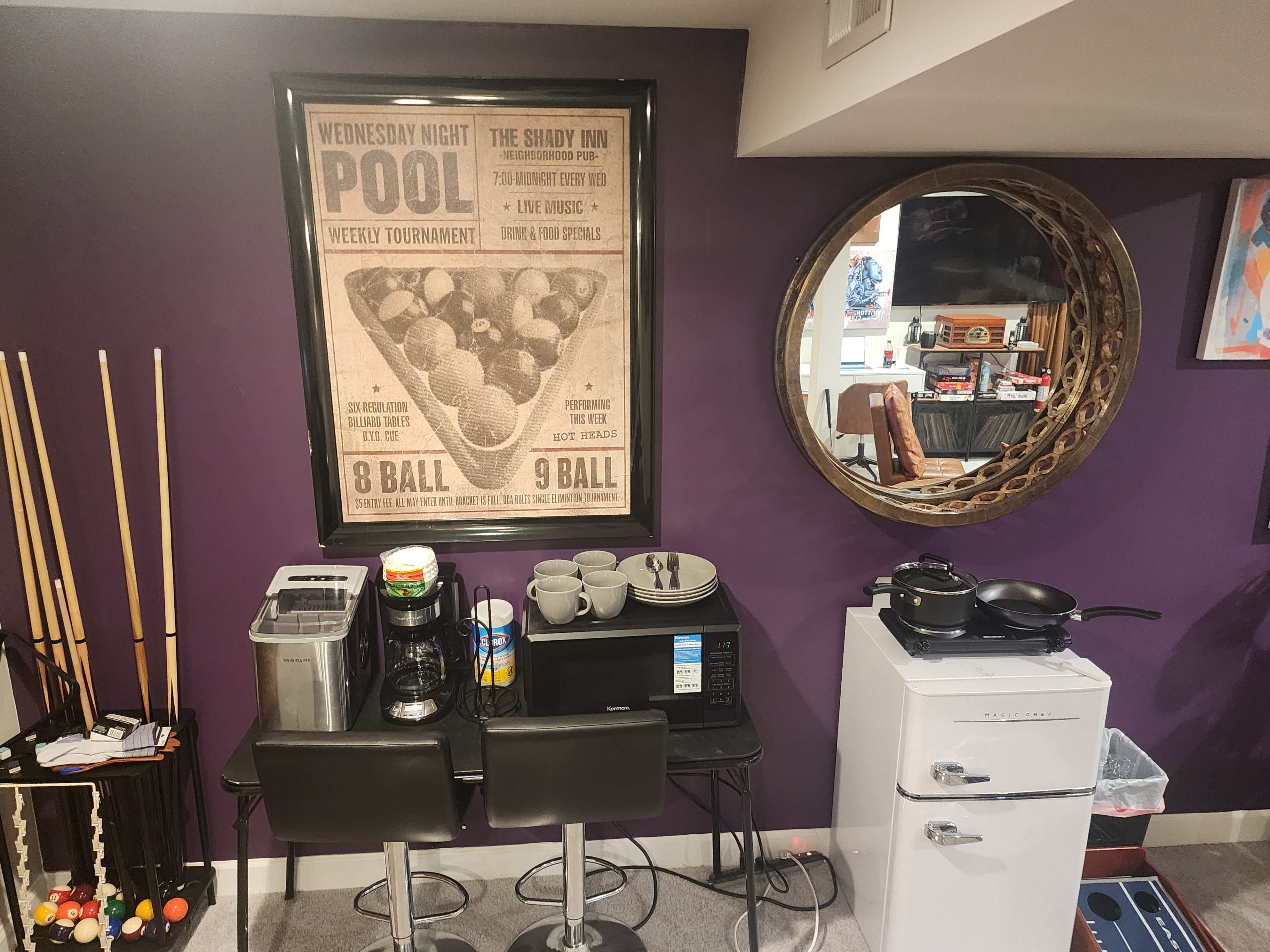 The image shows a small kitchenette area featuring a microwave, coffee maker, and a white fridge, with a framed vintage pool tournament poster on the wall.