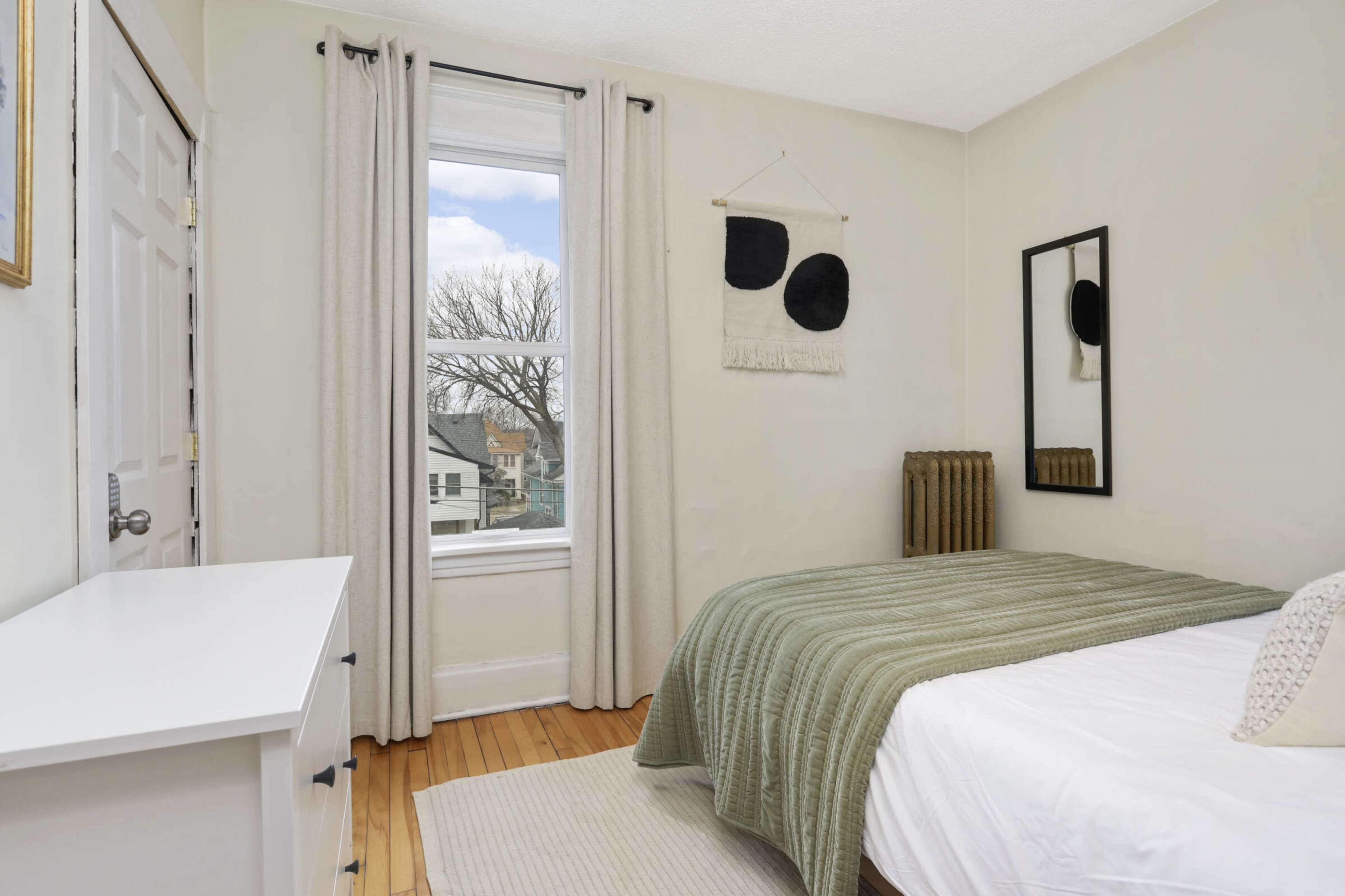 The image shows a simple bedroom with a bed covered in a green blanket, a mirror on the wall, and a window with curtains.