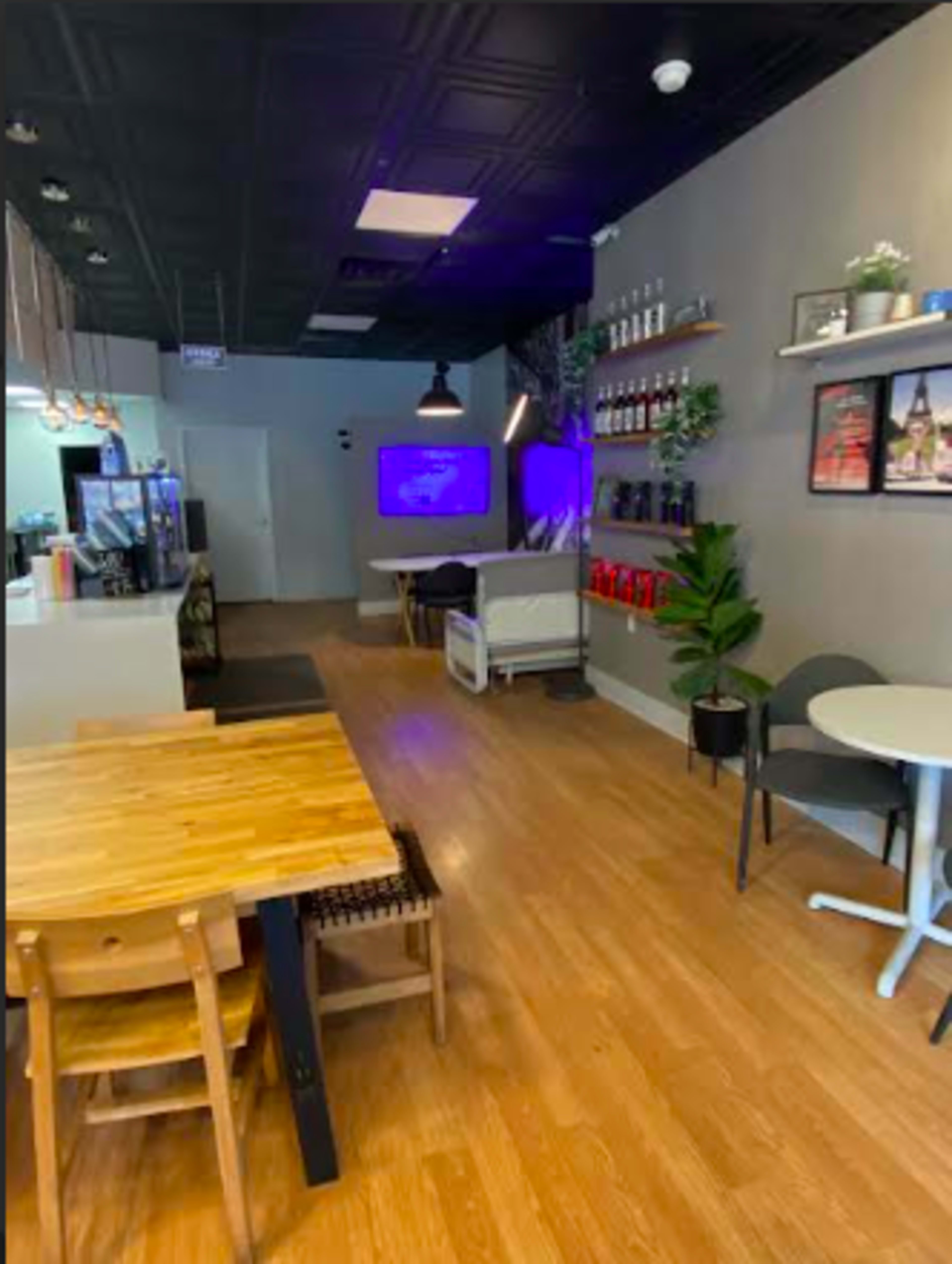 The image shows a cafe interior with wooden tables, chairs, and a wall-mounted screen displaying content, alongside a plant and decorative shelves.