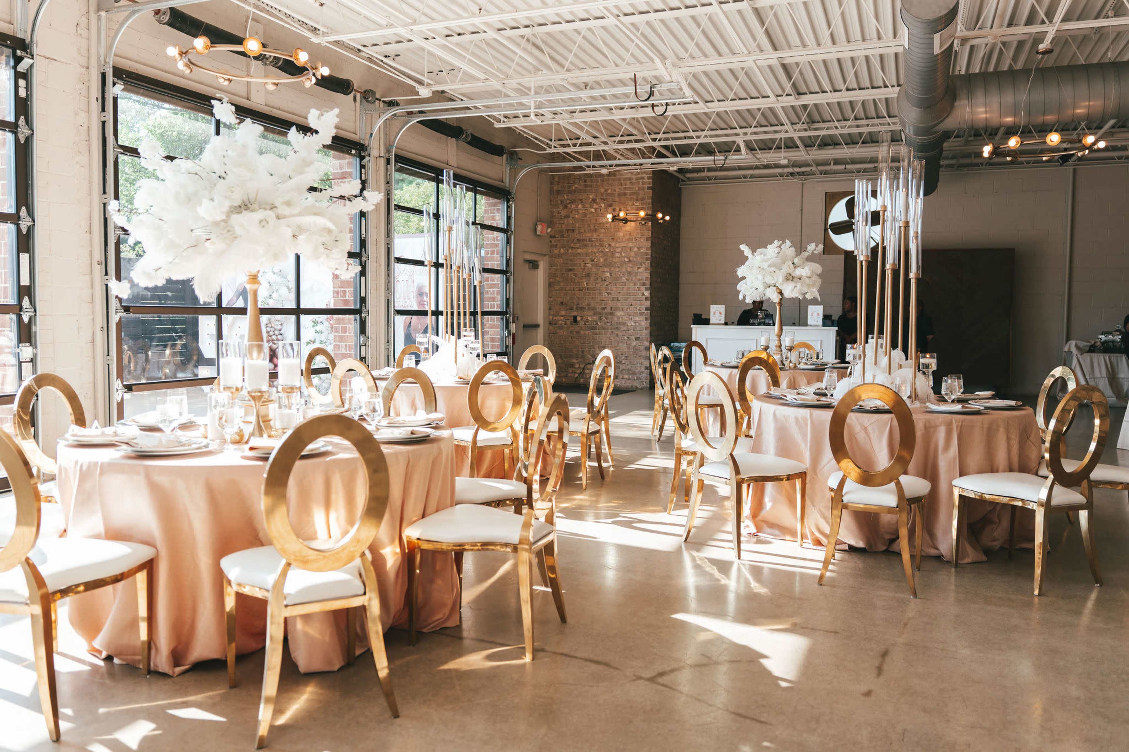 A spacious banquet hall features elegantly set tables with gold accents and floral centerpieces, illuminated by natural light streaming through large windows.