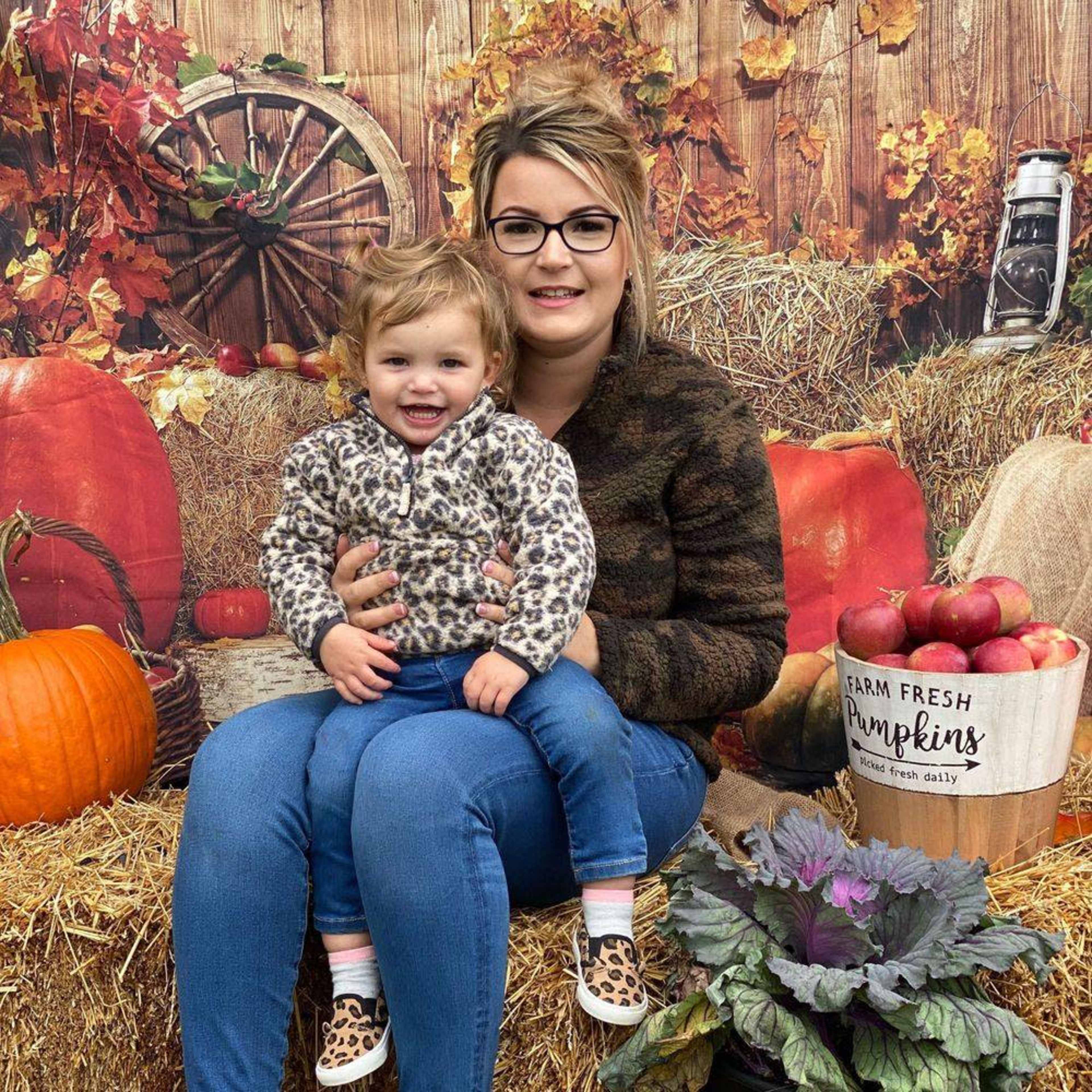 A woman and a young girl sit on a hay bale surrounded by pumpkins and autumn-themed decorations.