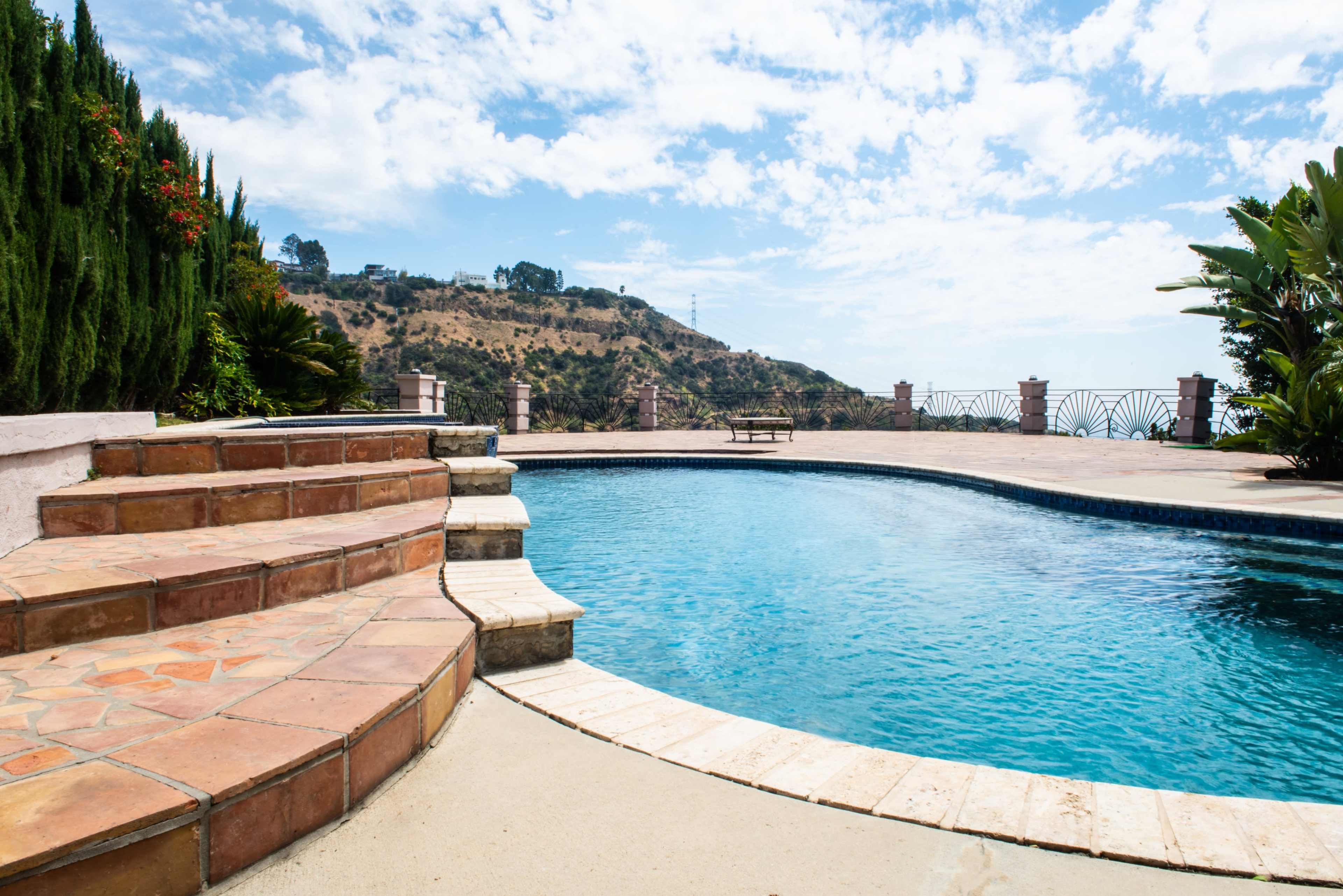 The image depicts a swimming pool with terracotta stone steps and landscaped greenery, set against a hilly backdrop under a cloudy sky.