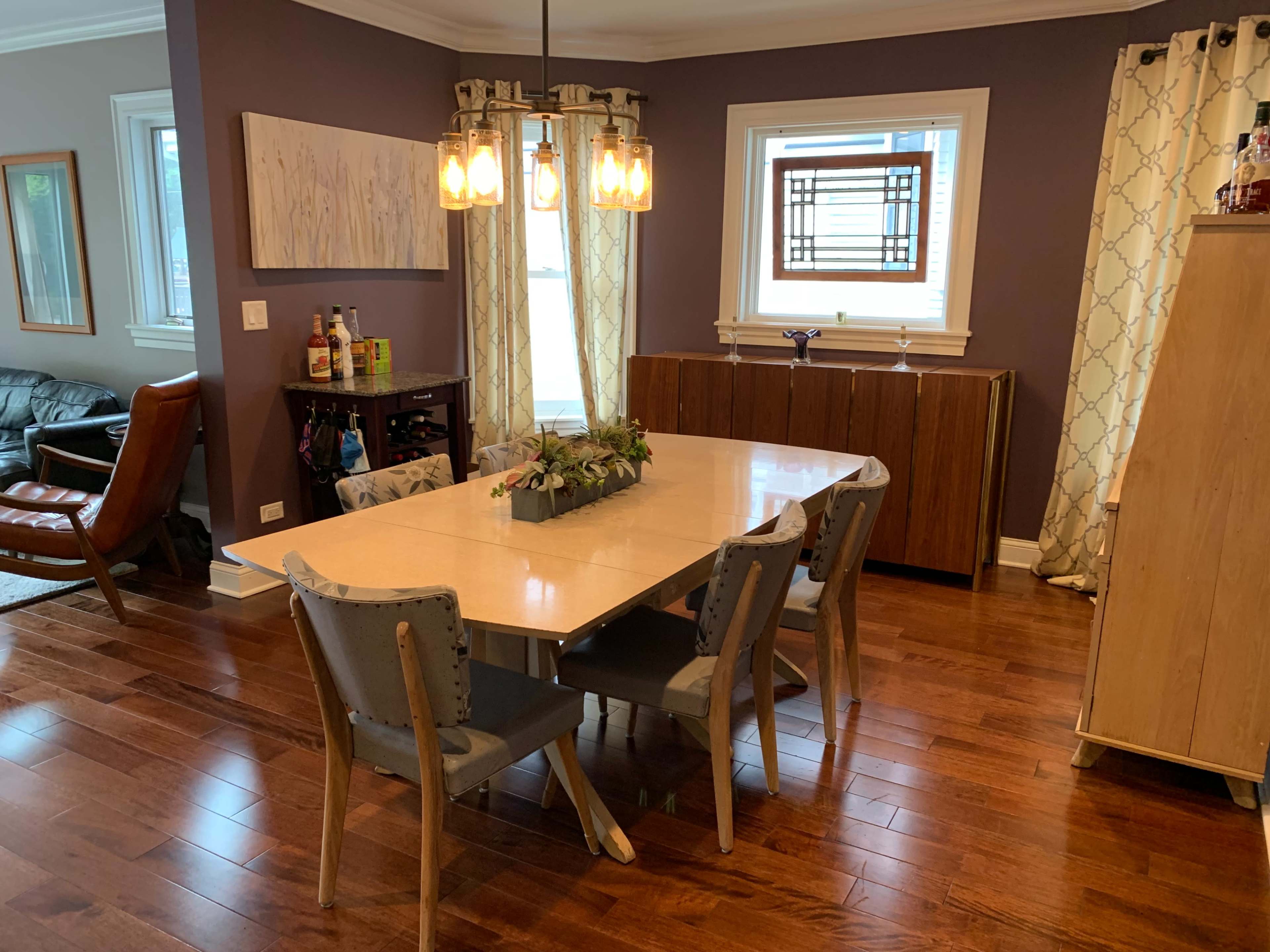 A dining area with a rectangular table surrounded by four chairs, a light fixture with hanging bulbs above, and a sideboard against a purple wall.