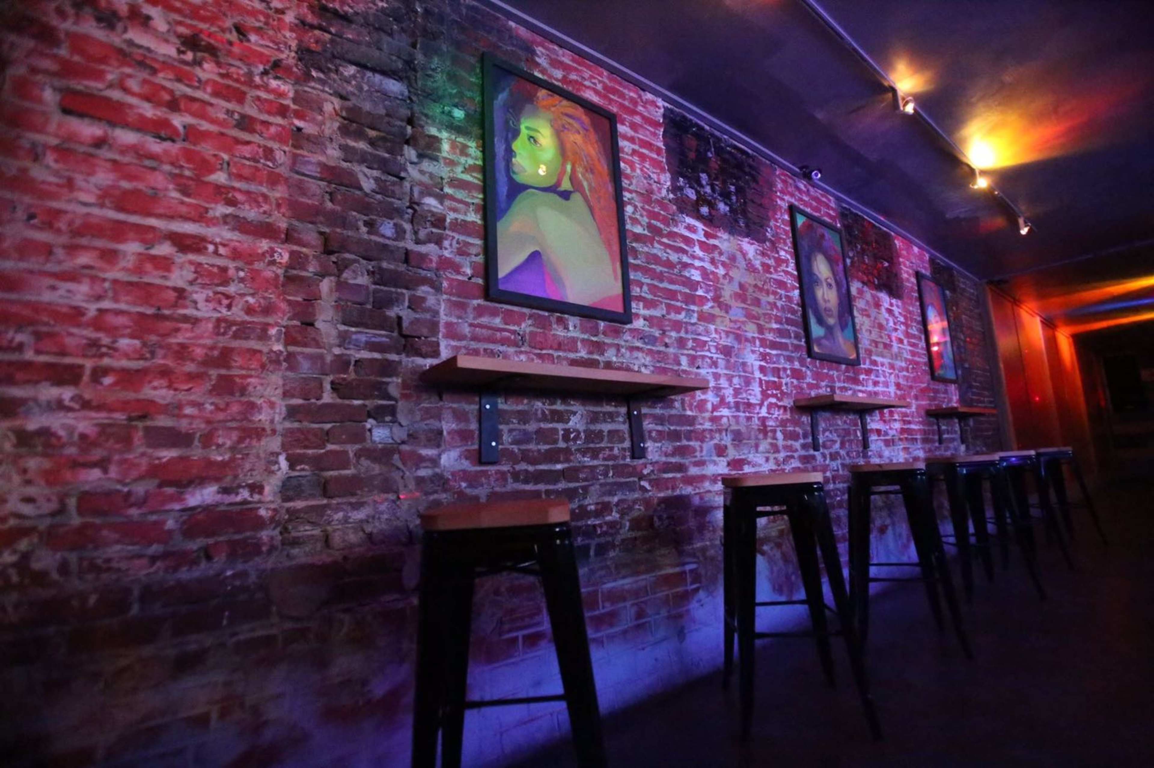 A dimly lit bar area with a brick wall adorned with framed portraits and a row of high stools along a wooden shelf.