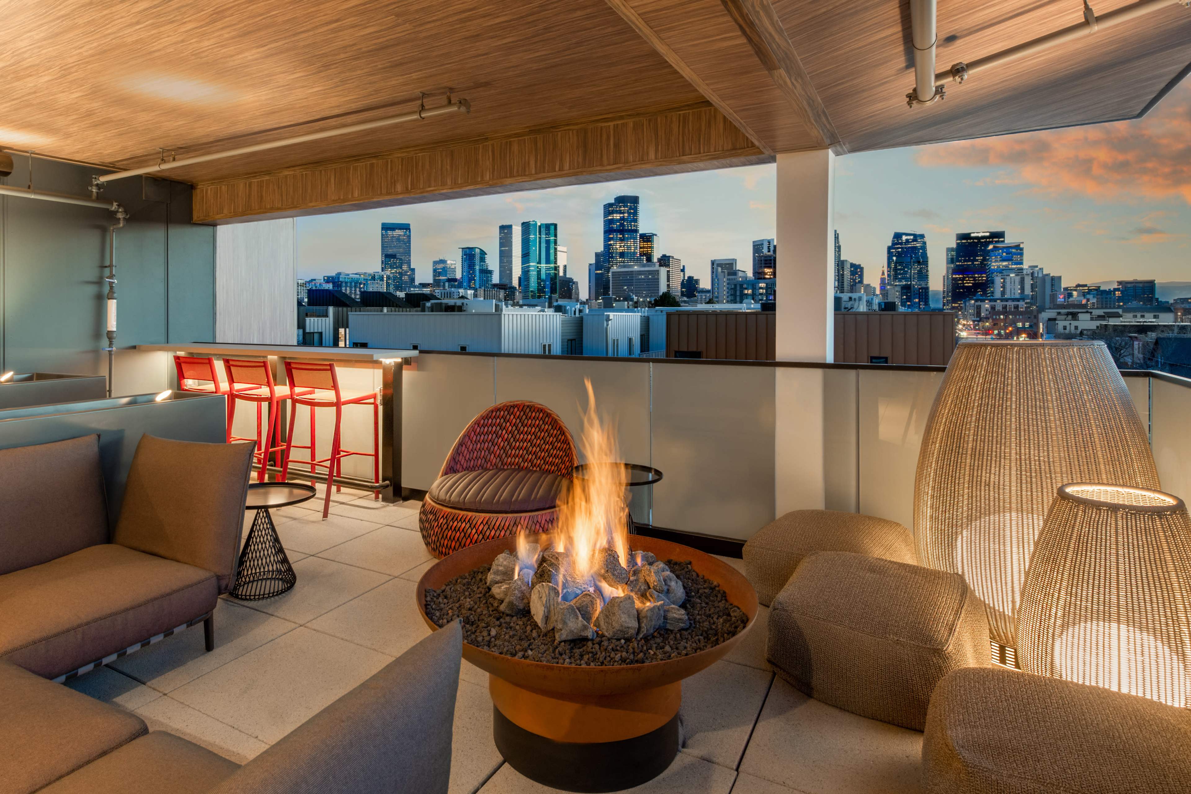 A modern rooftop lounge features a circular fire pit surrounded by comfortable seating, with a skyline view of tall buildings in the background at dusk.