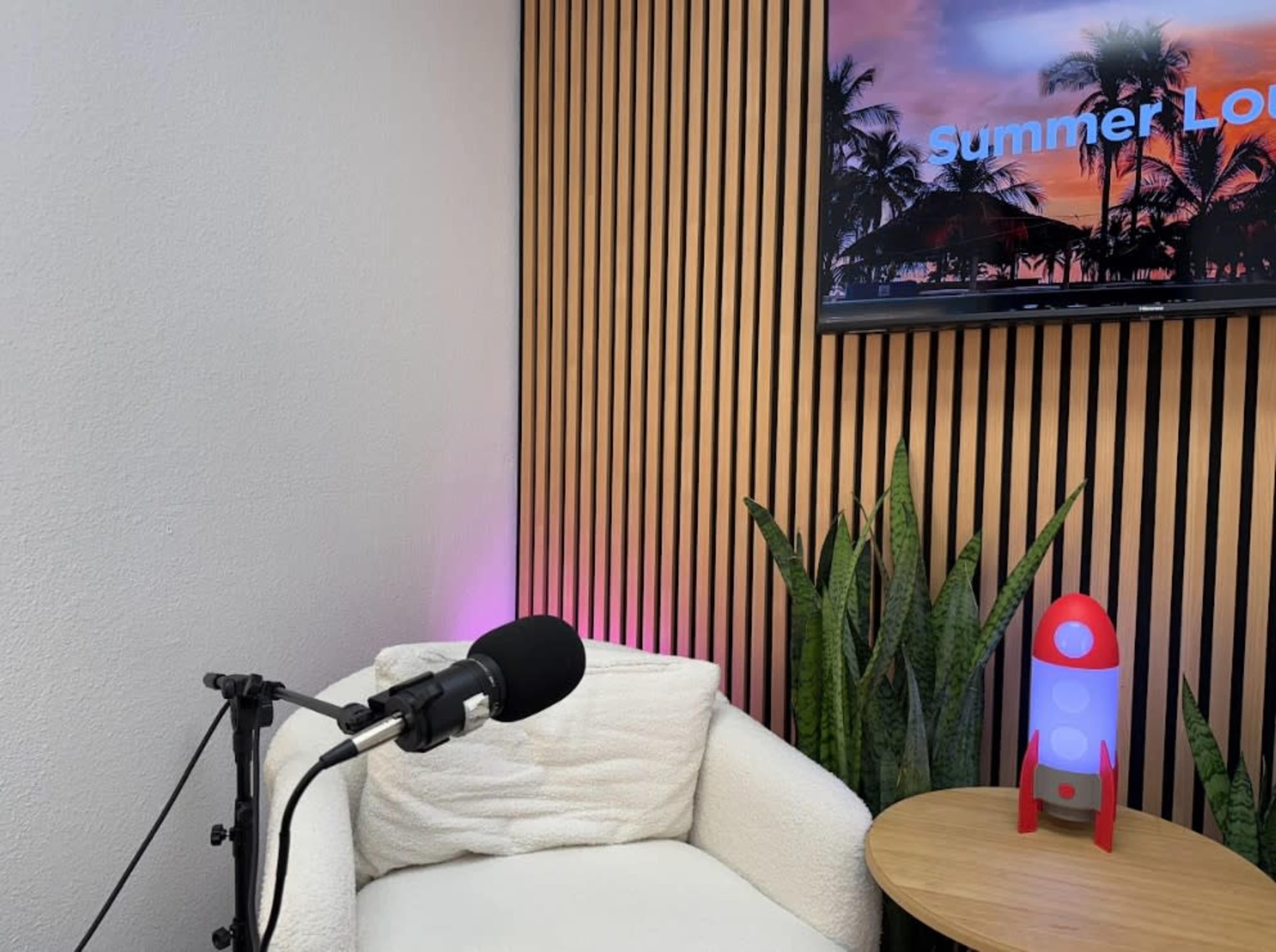 The scene features a cozy white chair next to a microphone and a small table, with a colorful backdrop displaying a summer-themed image and decorative plants.