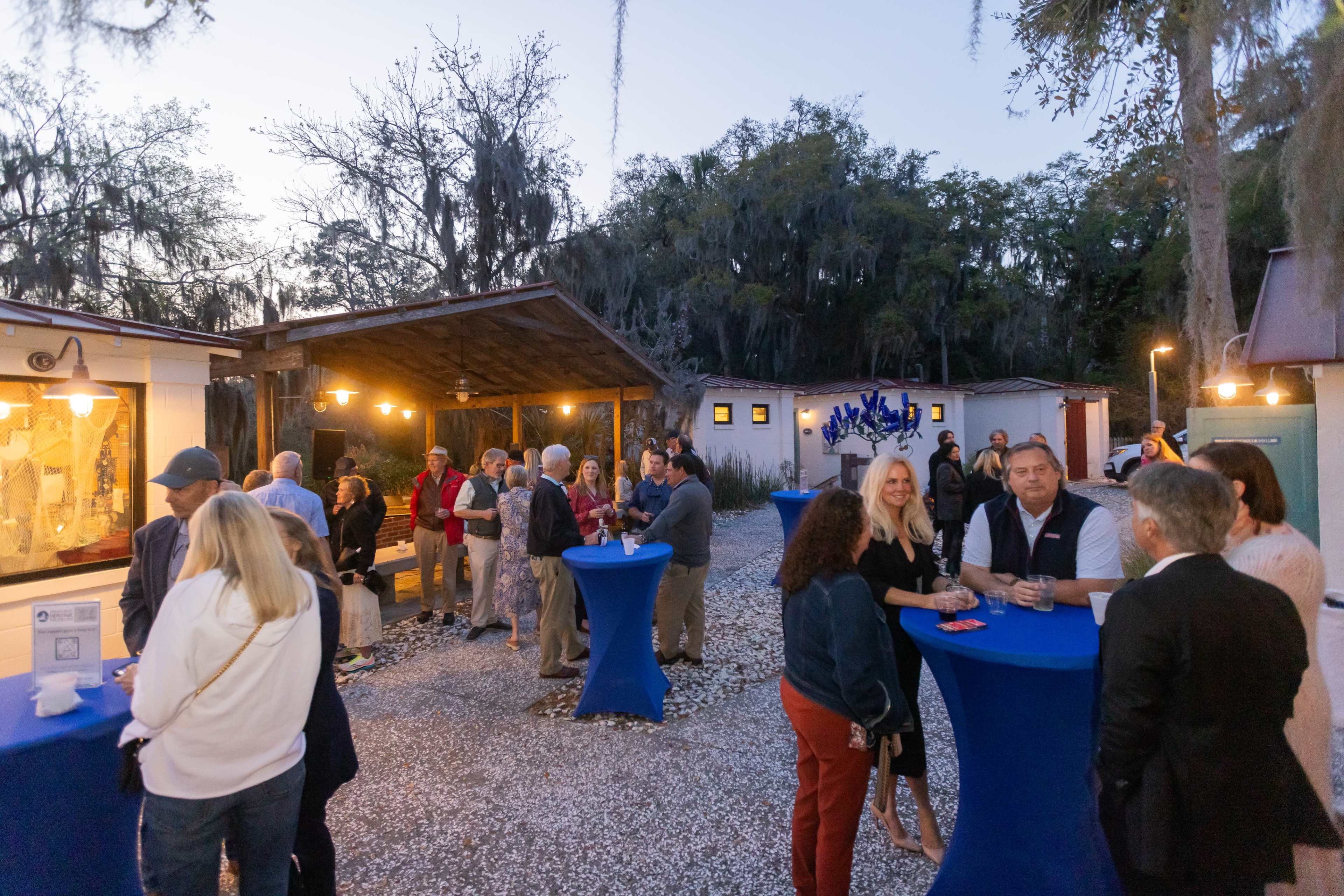 A group of people gathered around blue tables outside in a landscaped area with trees and rustic buildings during twilight.