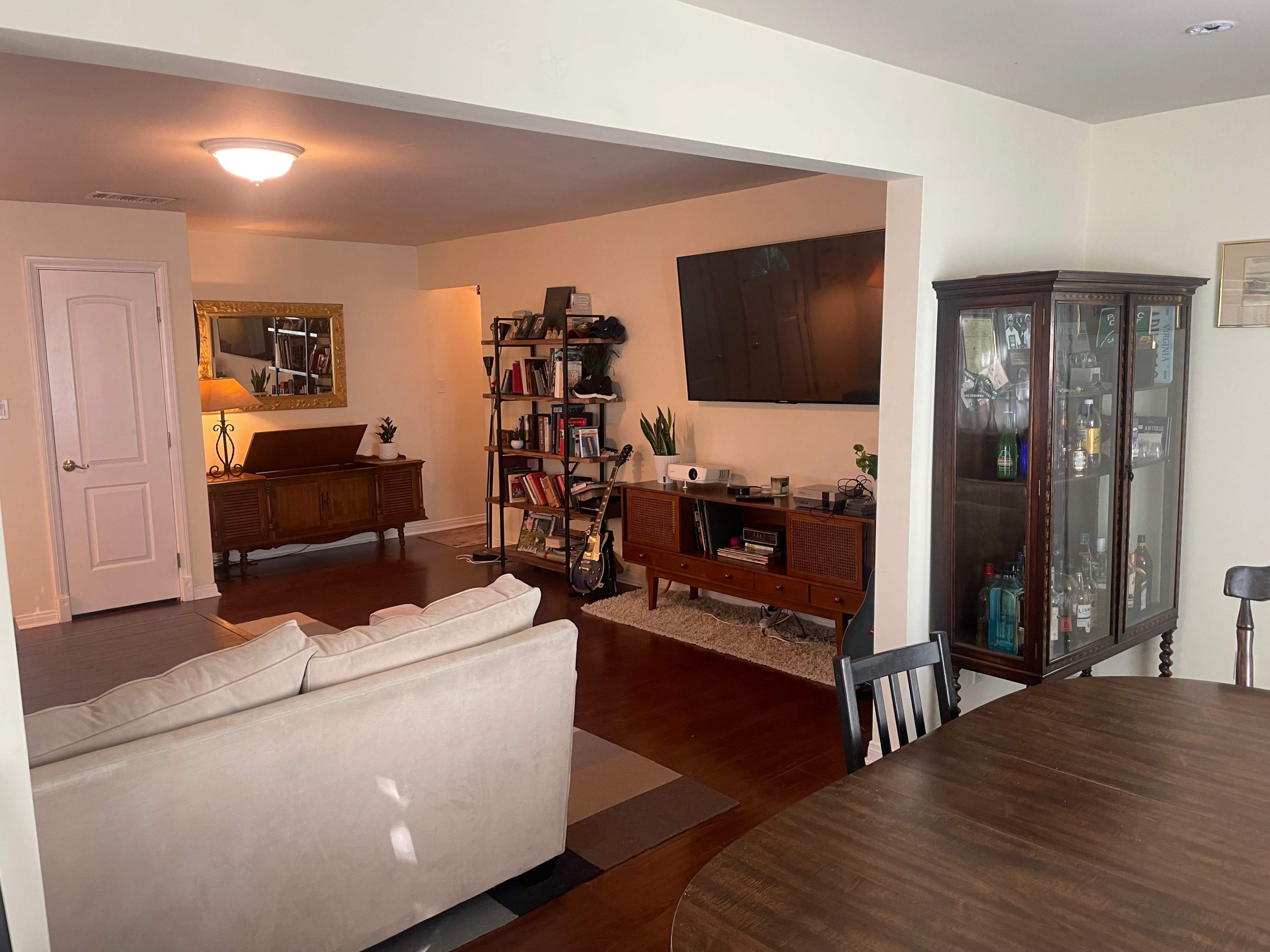 The image shows a living space with a sectional sofa, a dining area, a large TV mounted on the wall, bookshelves, and a cabinet displaying various items.