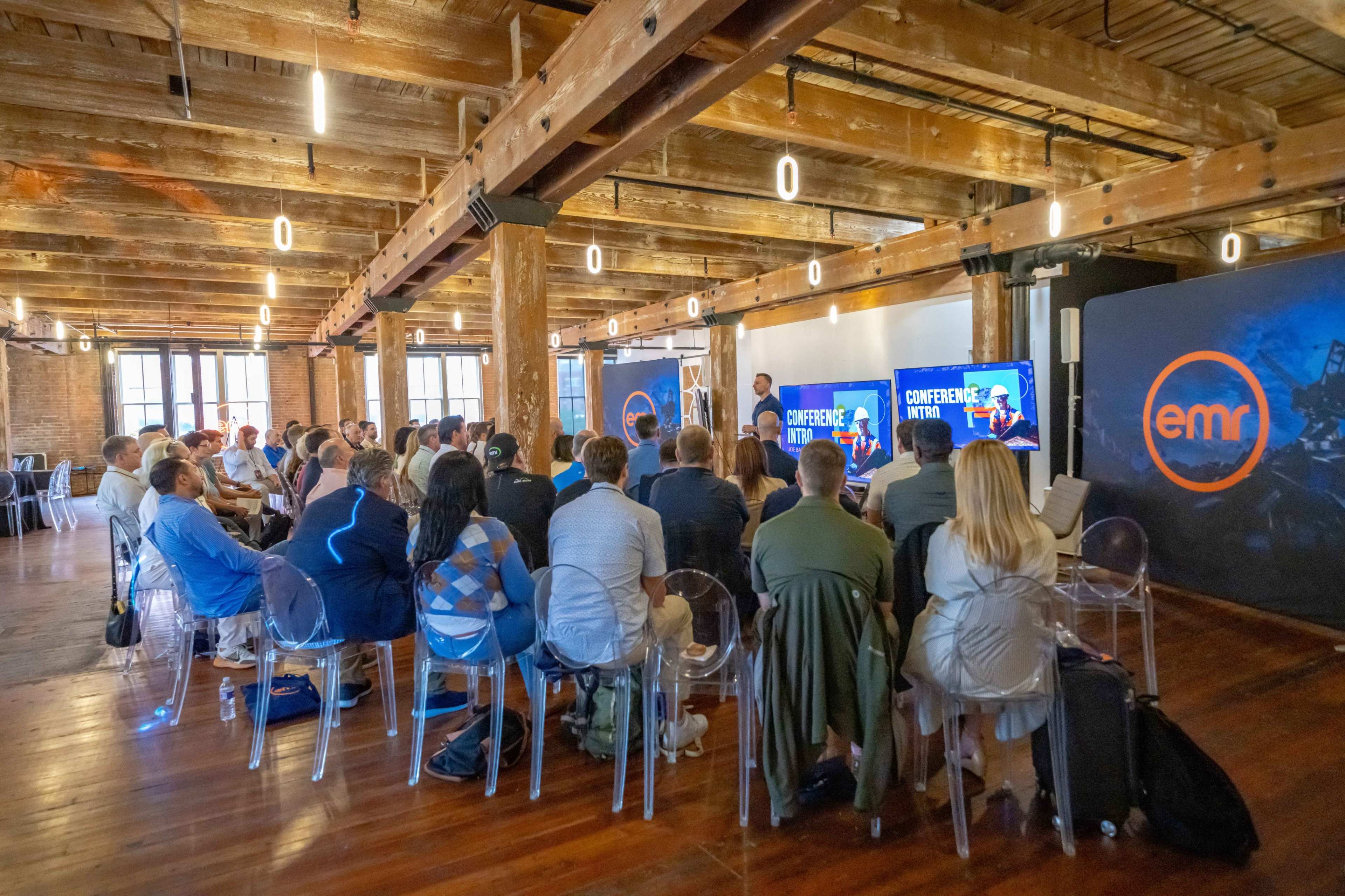 A speaker presents to an audience in a spacious, brightly lit conference room with wooden beams and large screens displaying the event's title.