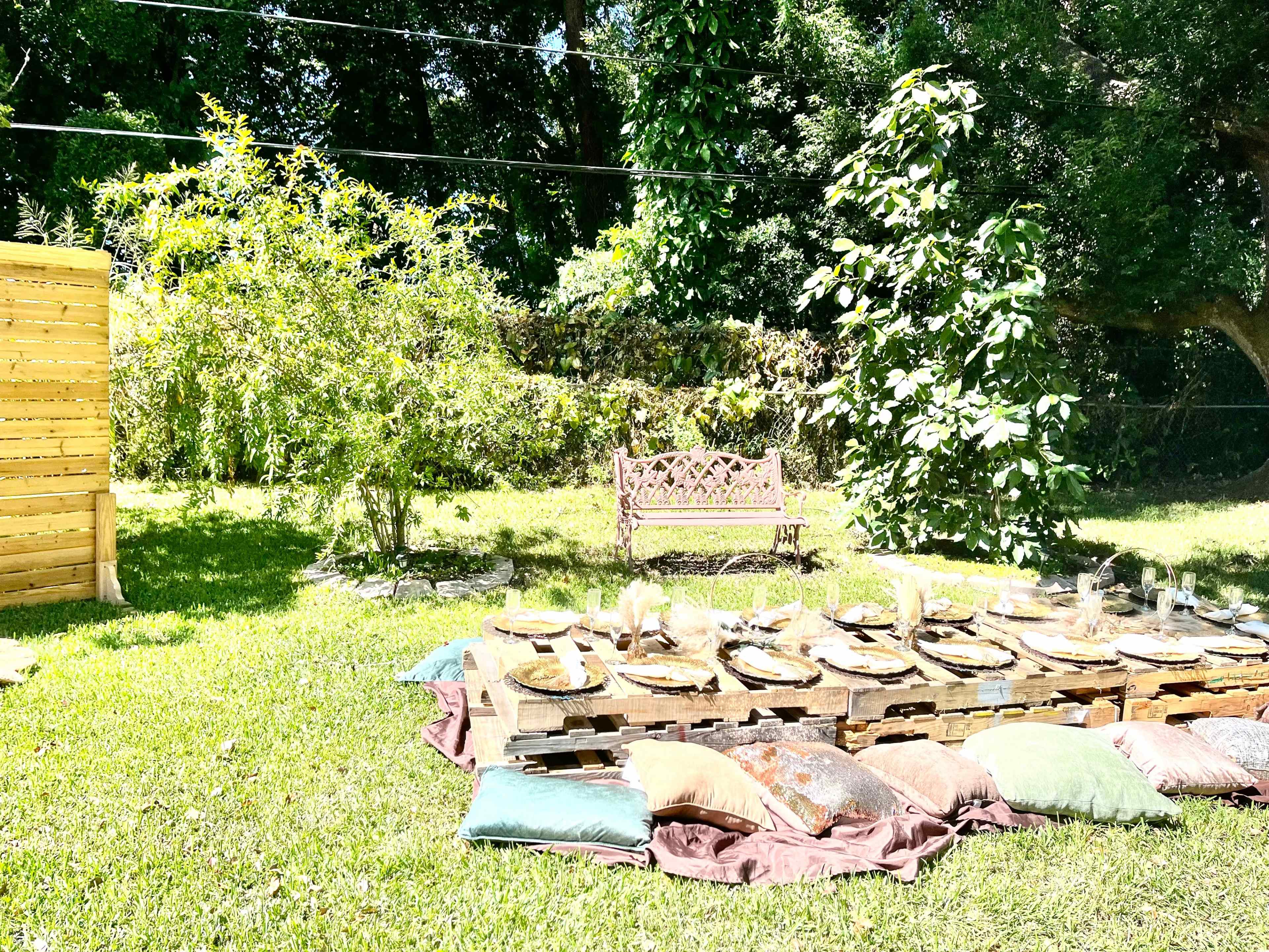 A picnic setup with wooden pallets as a table, decorated with plates and cushions, is arranged on green grass under trees.