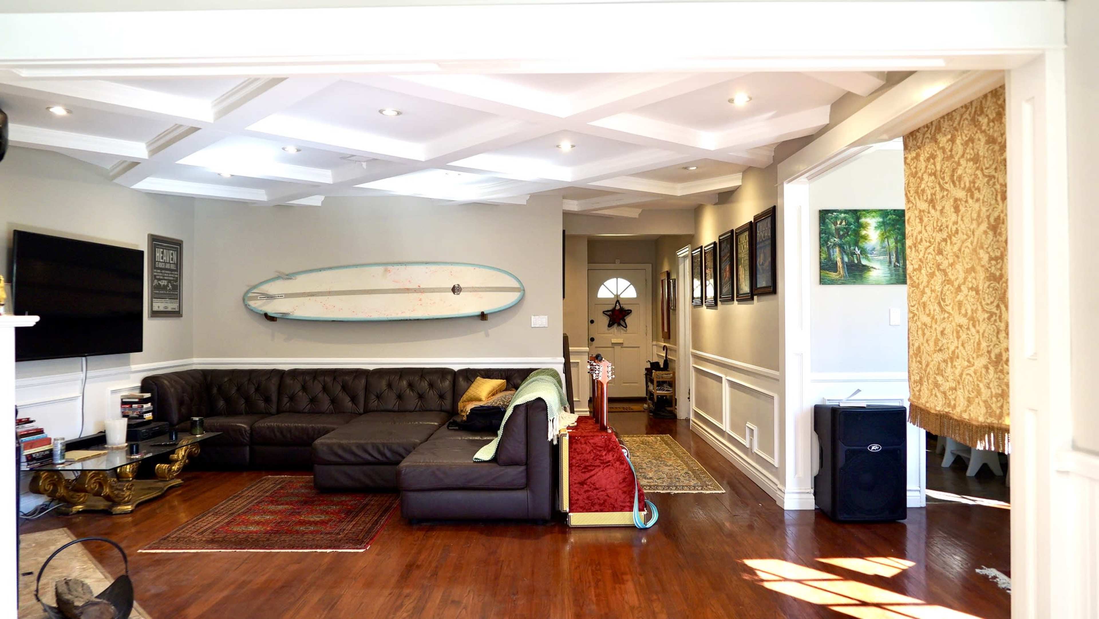 The image shows a spacious living room with a dark sectional couch, a long surfboard mounted on the wall, and a doorway leading to another room.