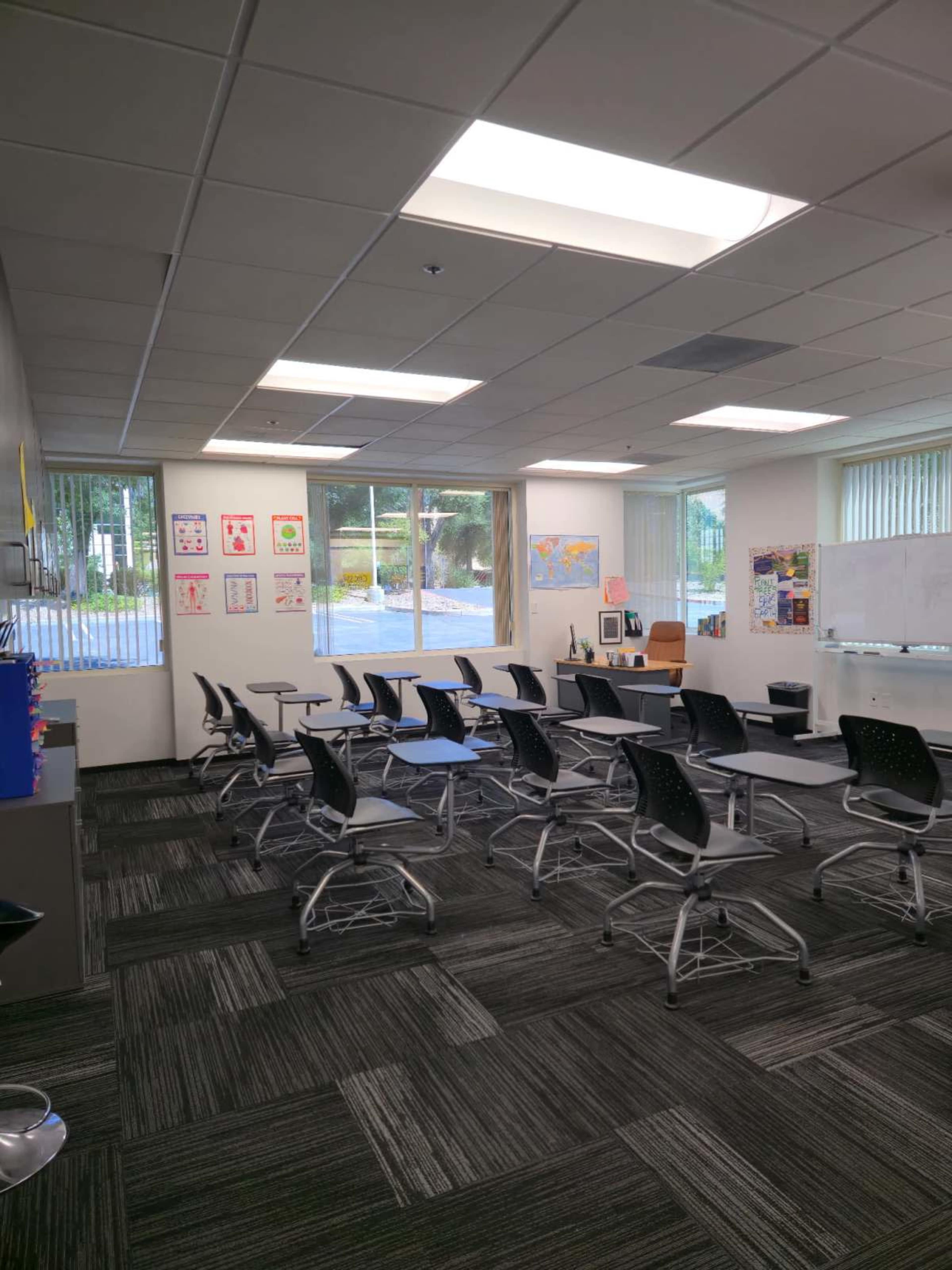 The image shows a classroom with rows of movable desks, large windows letting in natural light, and various educational posters on the walls.