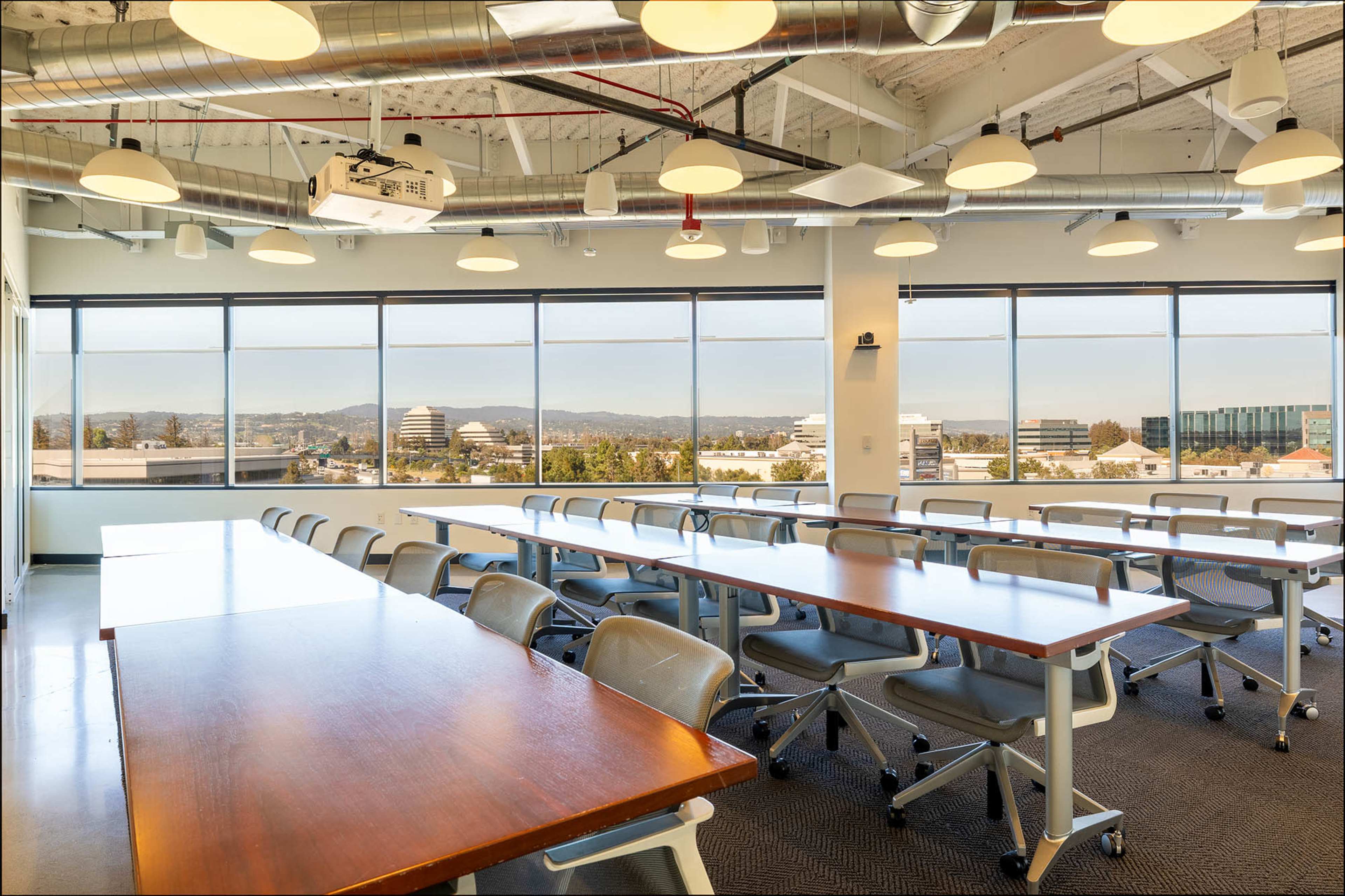 A spacious conference room with multiple tables arranged in rows, large windows providing a view of the landscape outside, and overhead lighting fixtures.