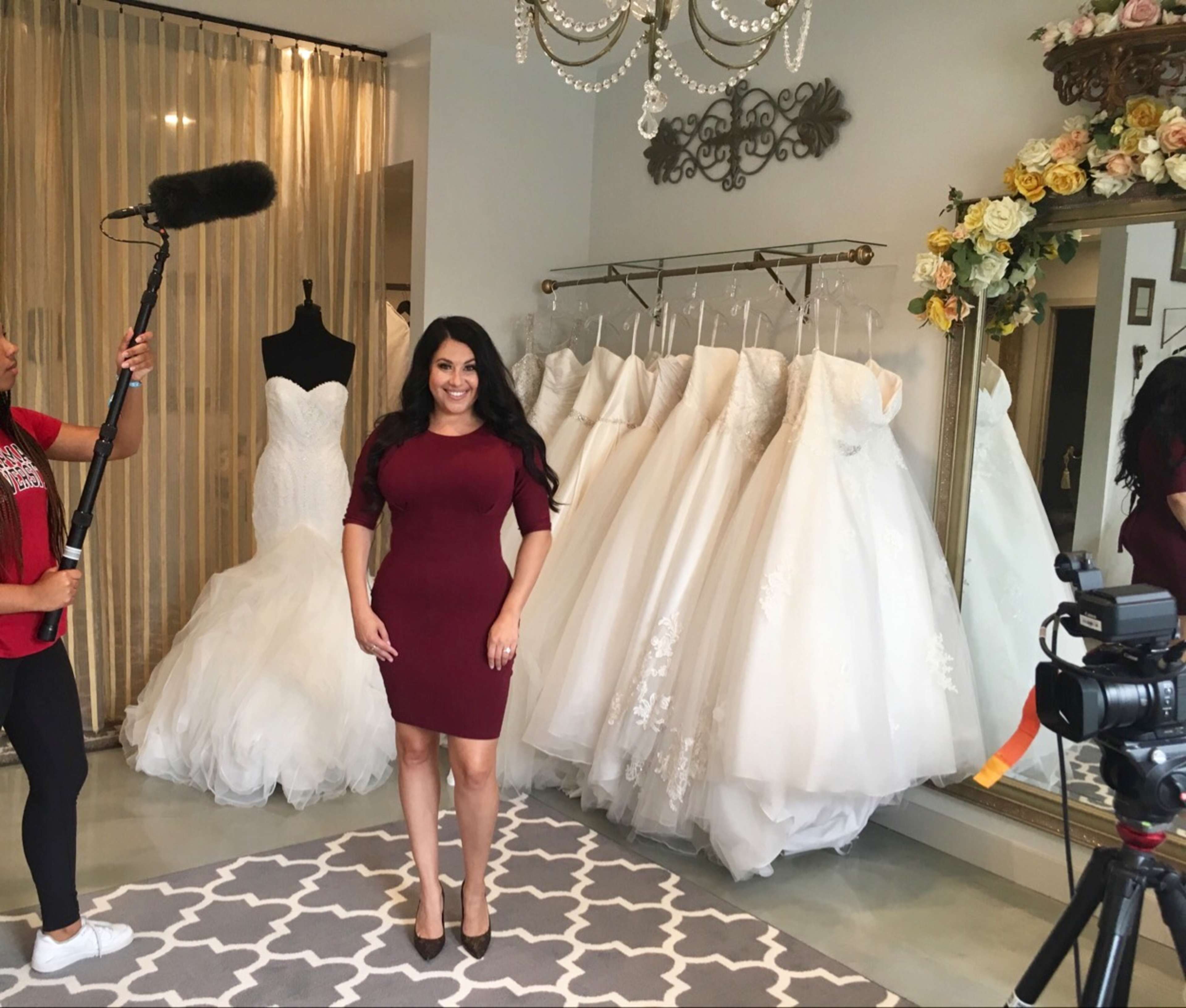 A woman stands in a bridal shop surrounded by white wedding dresses, with a microphone on a boom being held nearby and cameras set up for filming.