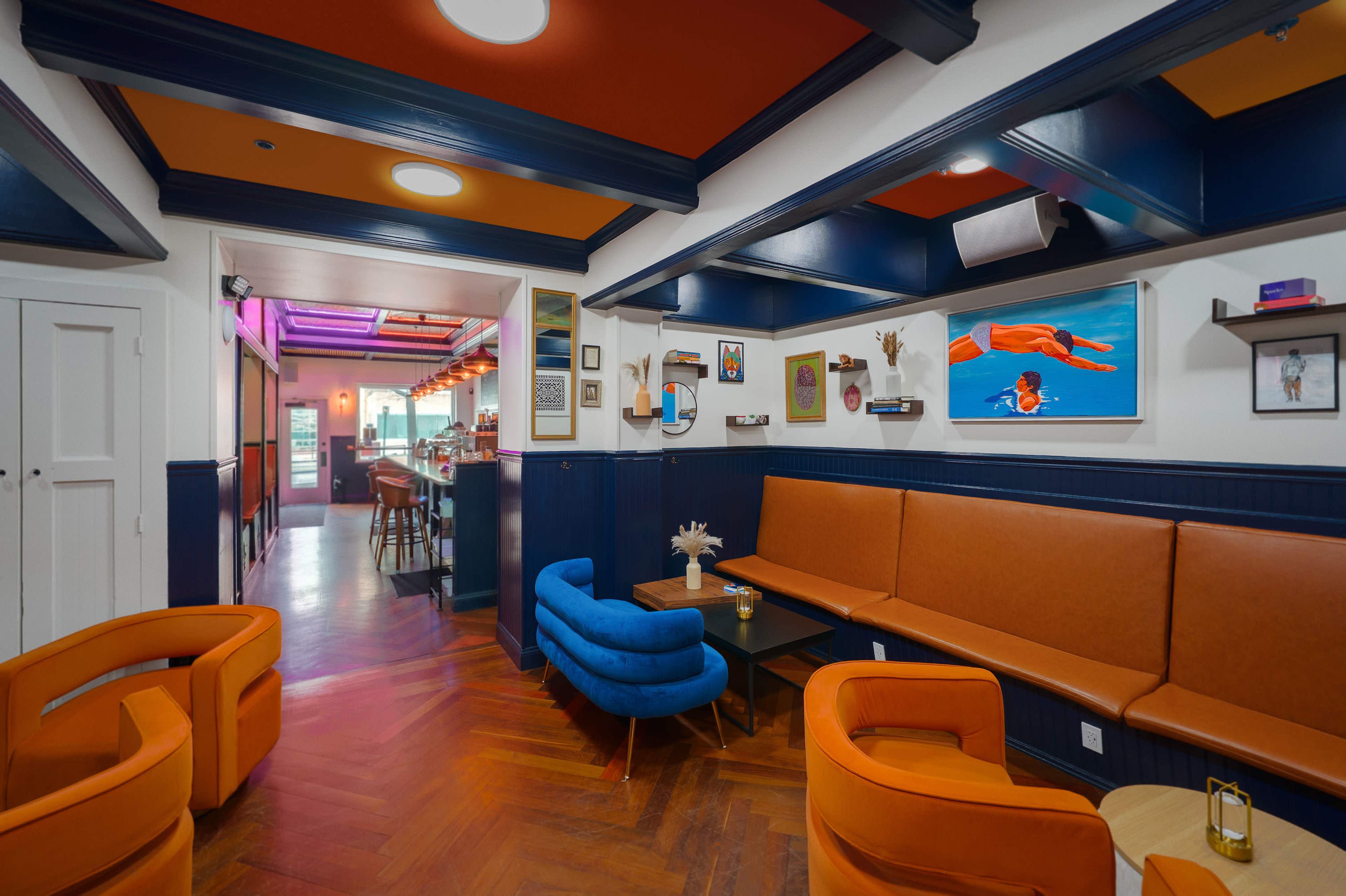 The image shows a brightly colored cafe interior featuring orange and blue seating, wooden flooring, and various artworks on the walls.