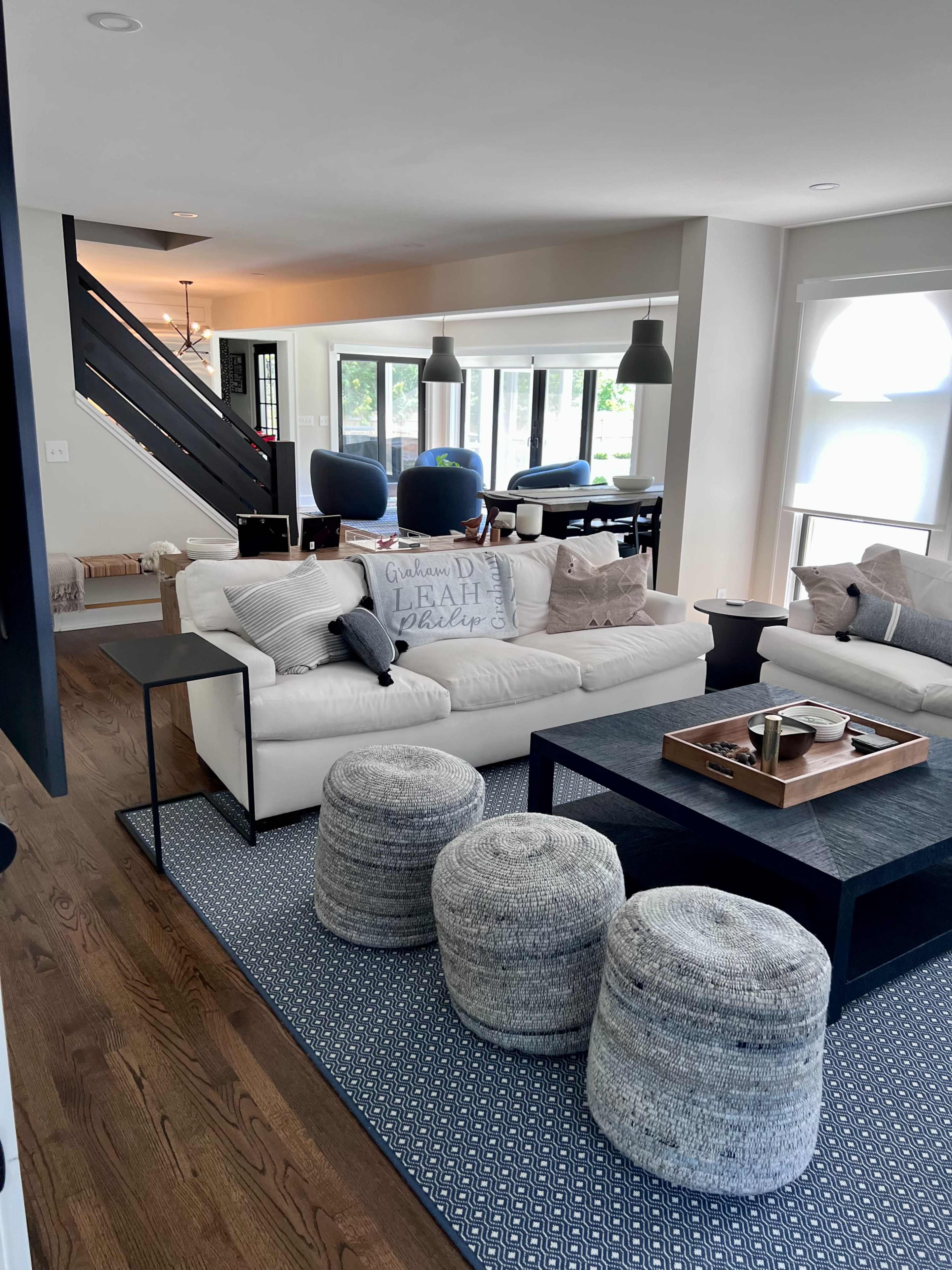 The image shows a modern living room featuring a white sectional sofa, a dark coffee table, and a light-colored area rug, with a dining area visible in the background.