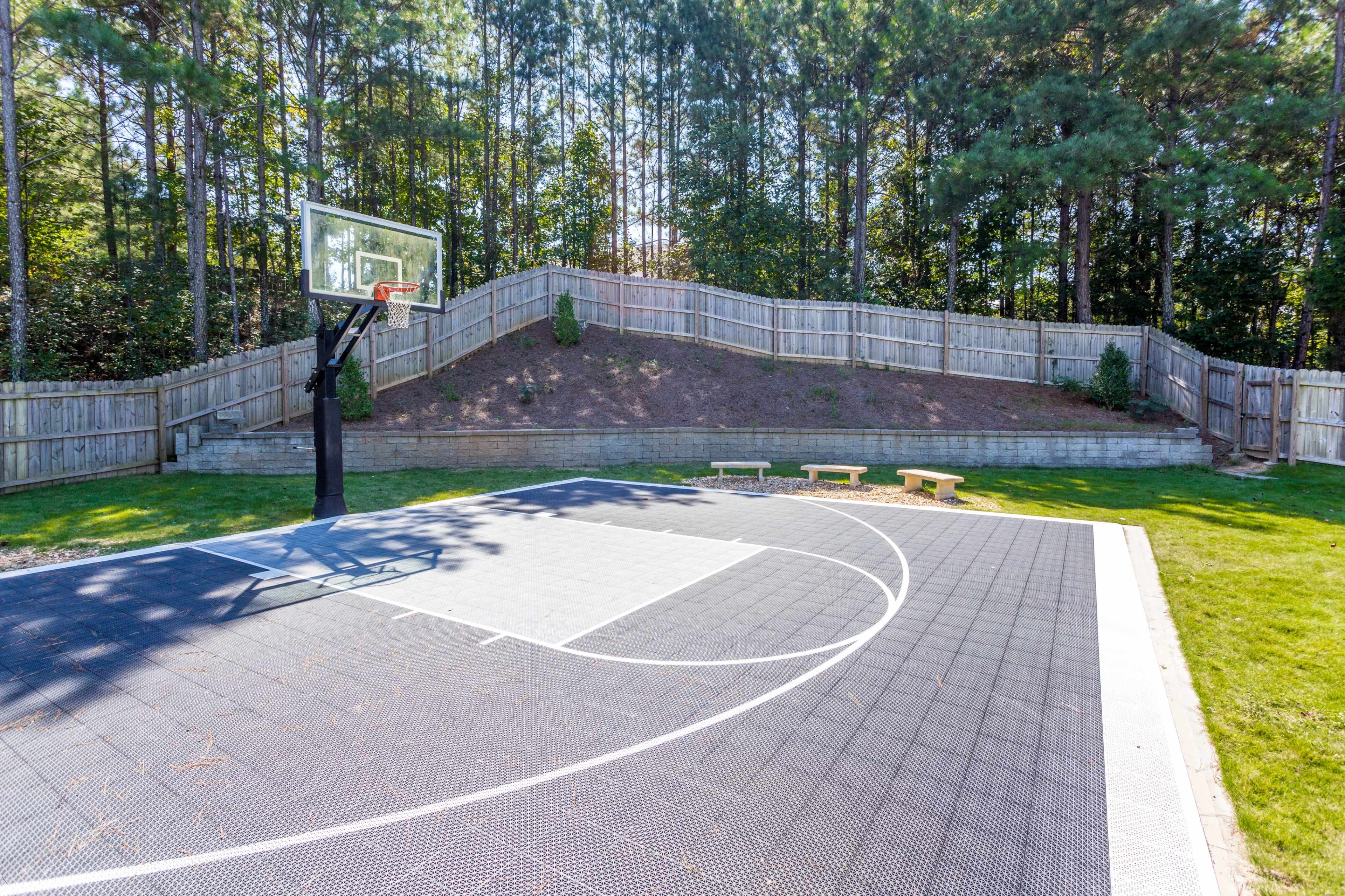 A backyard basketball court with a hoop, surrounded by trees and a wooden fence.