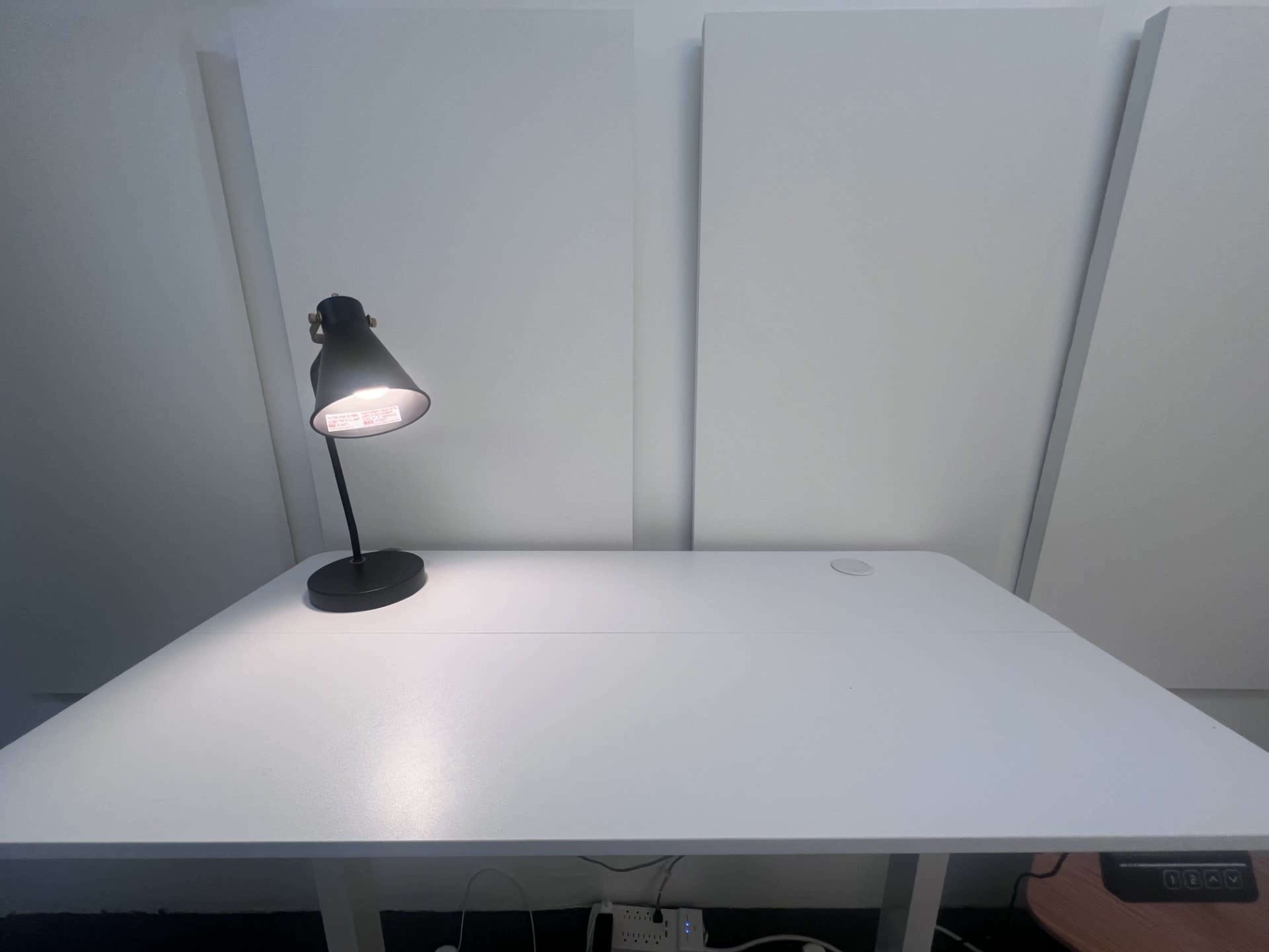 A white desk with a black lamp is positioned against a backdrop of three white panels.