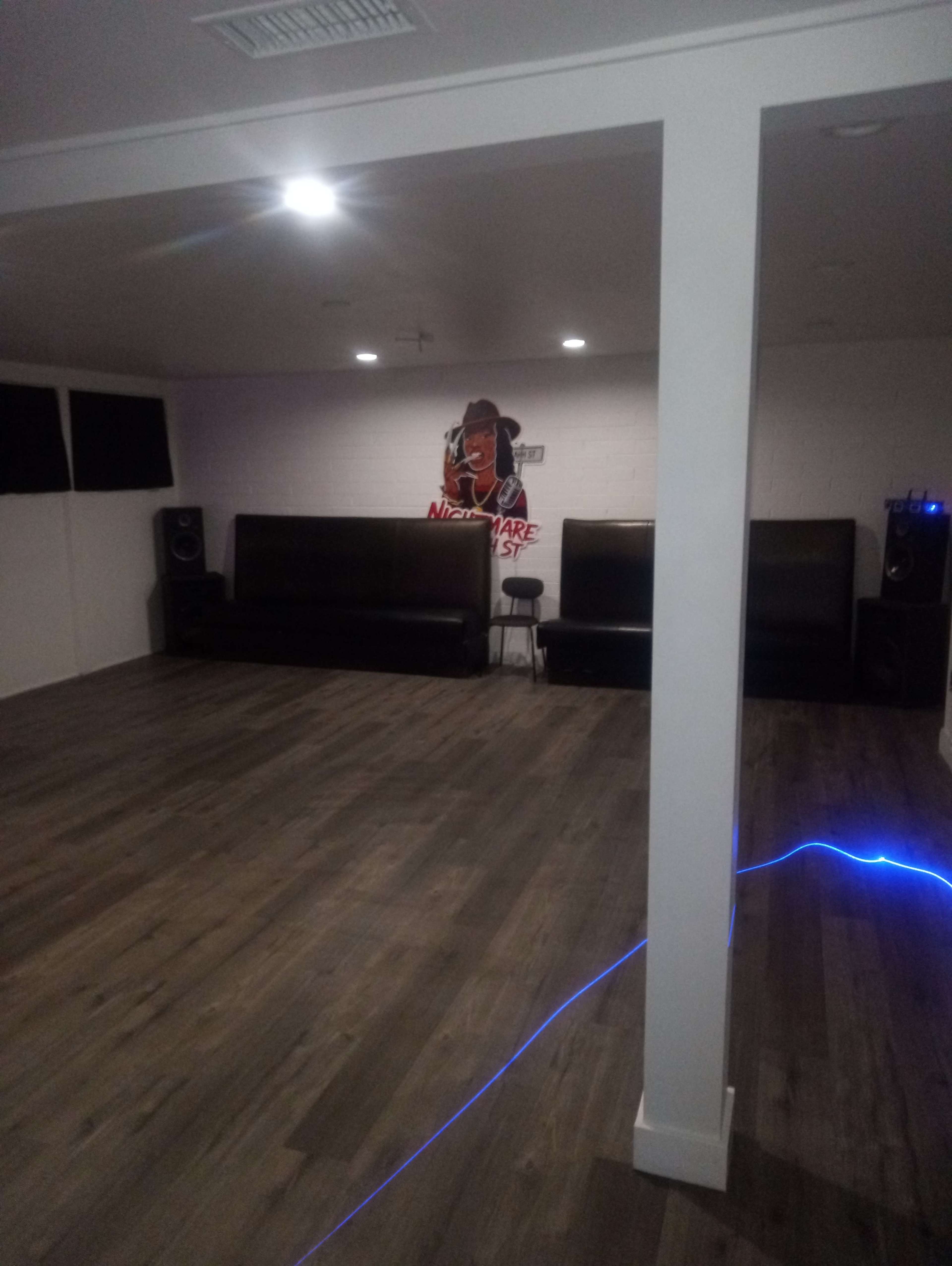 The image shows a spacious room with wooden flooring, two dark leather couches facing a wall featuring a mural of a character, and speakers placed on either side.