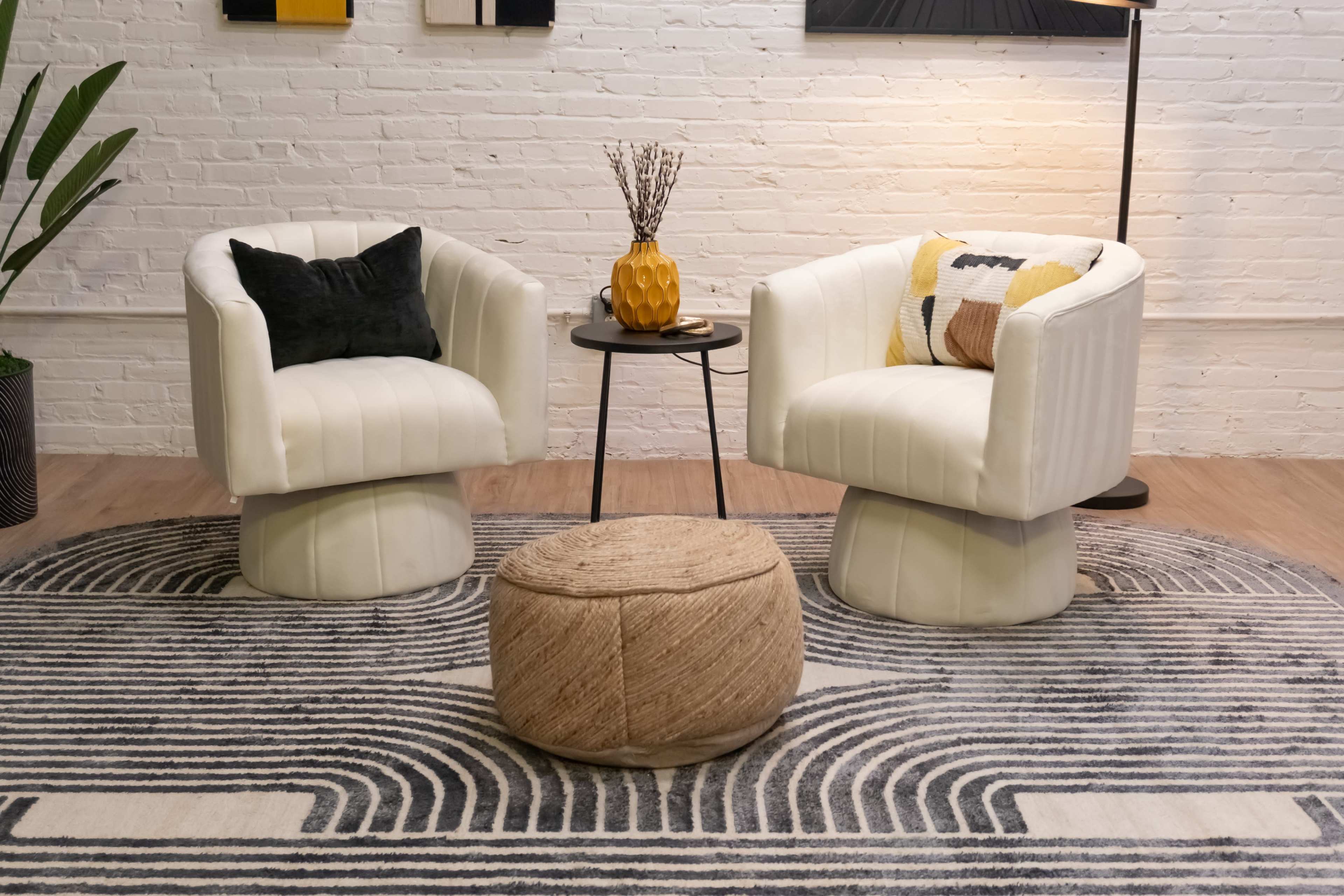 The image shows a cozy seating area with two white armchairs, a small black side table, a round woven ottoman, and a patterned rug on the floor.