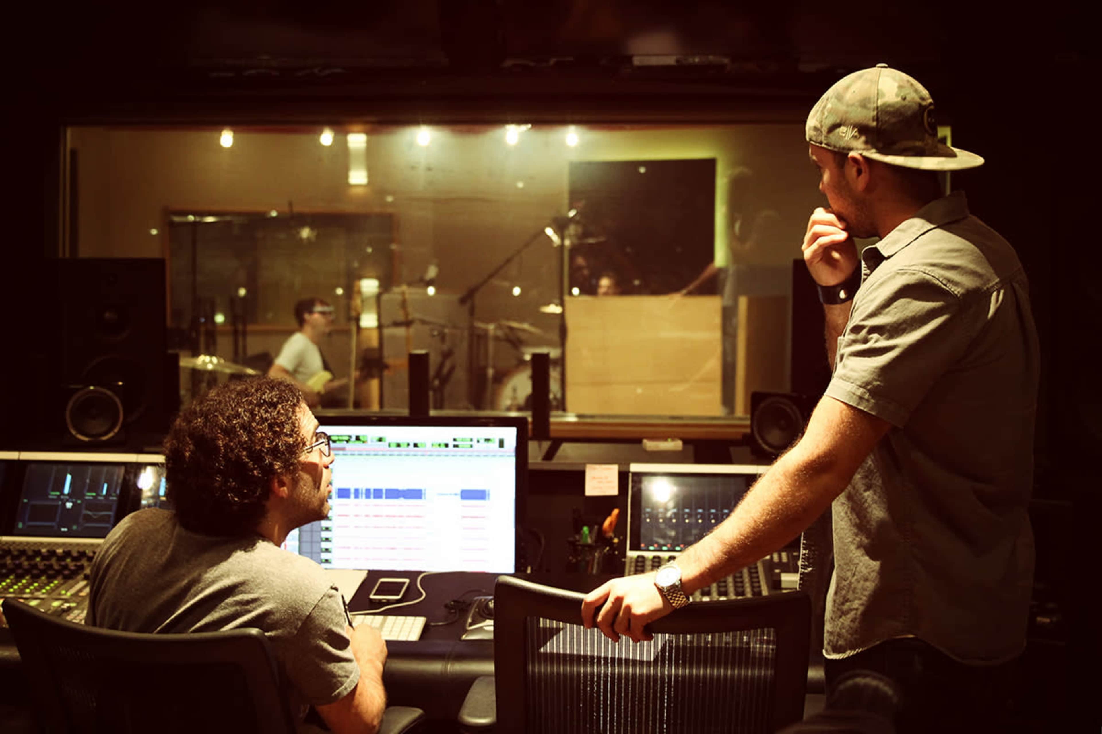 Two men are working in a music recording studio, one seated at a control desk while the other stands nearby, observing the recording session through a glass partition.