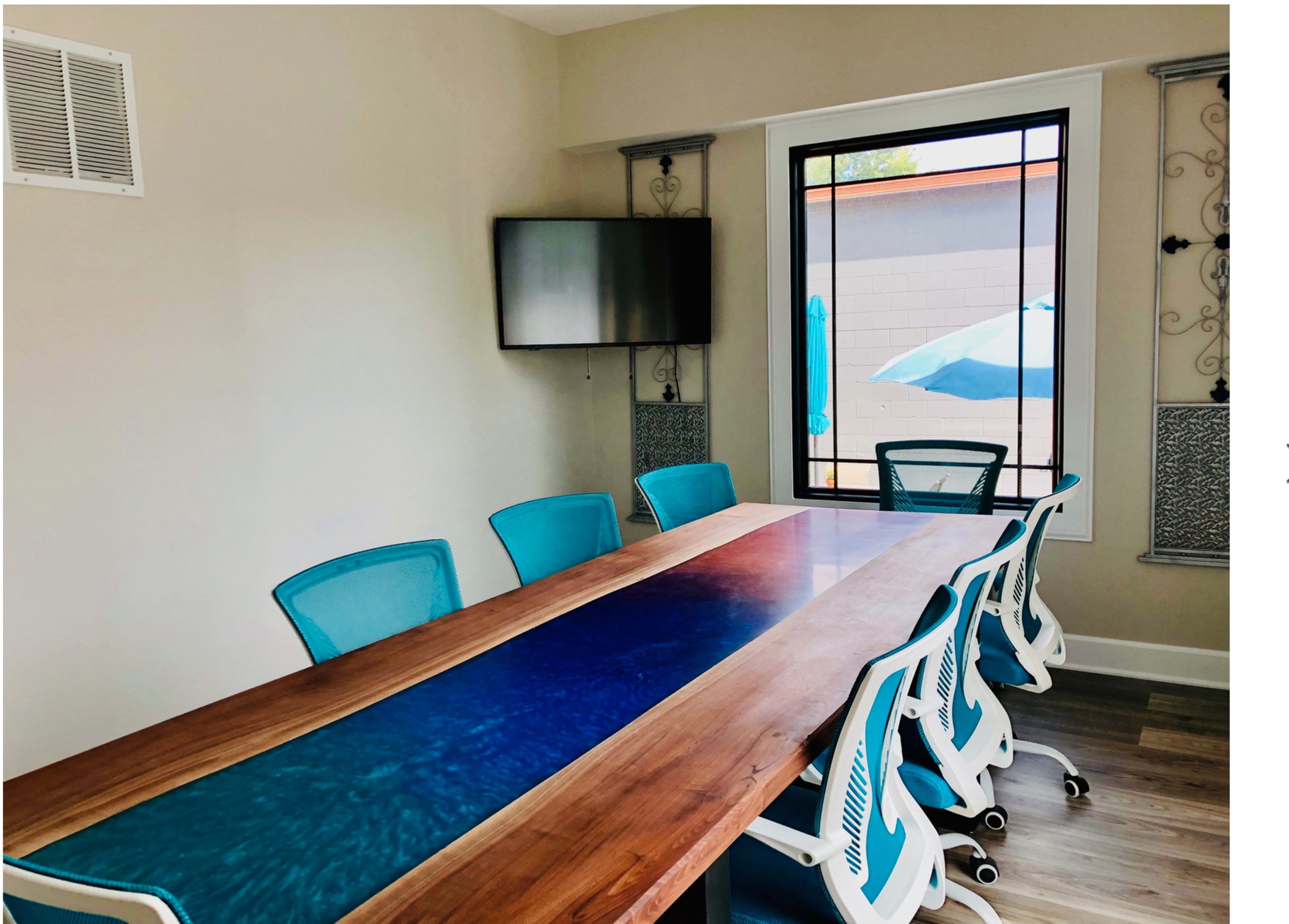 The image shows a conference room featuring a large wooden table with a blue resin inlay and six blue chairs arranged around it, along with a TV mounted on the wall.