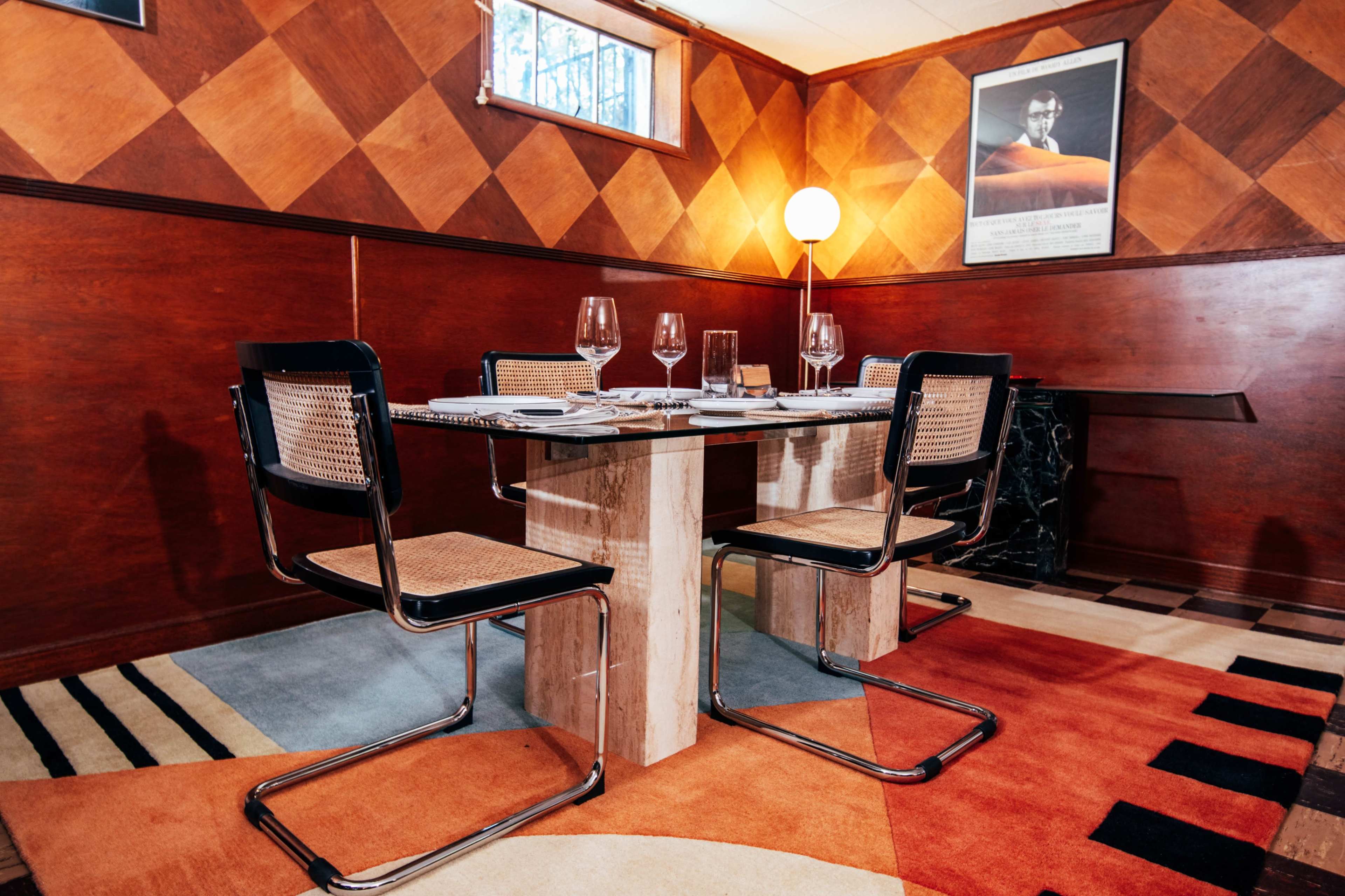 A dining area features a square table with a marble base surrounded by four modern chairs, set against a backdrop of wooden paneling and geometric-patterned carpets.