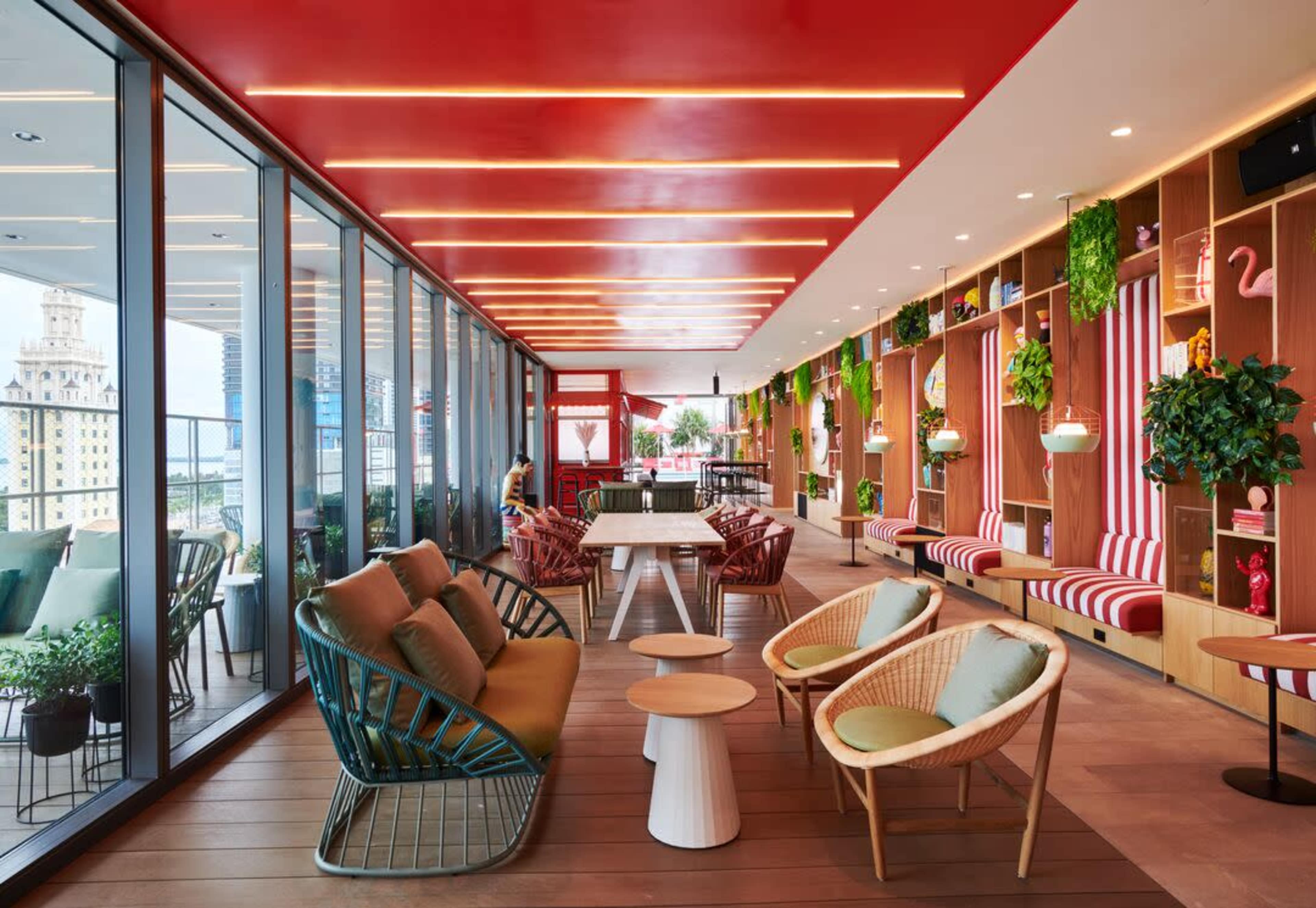 A modern indoor lounge with a vibrant red ceiling, large windows, and a mix of comfortable seating surrounded by greenery and decorative shelving.