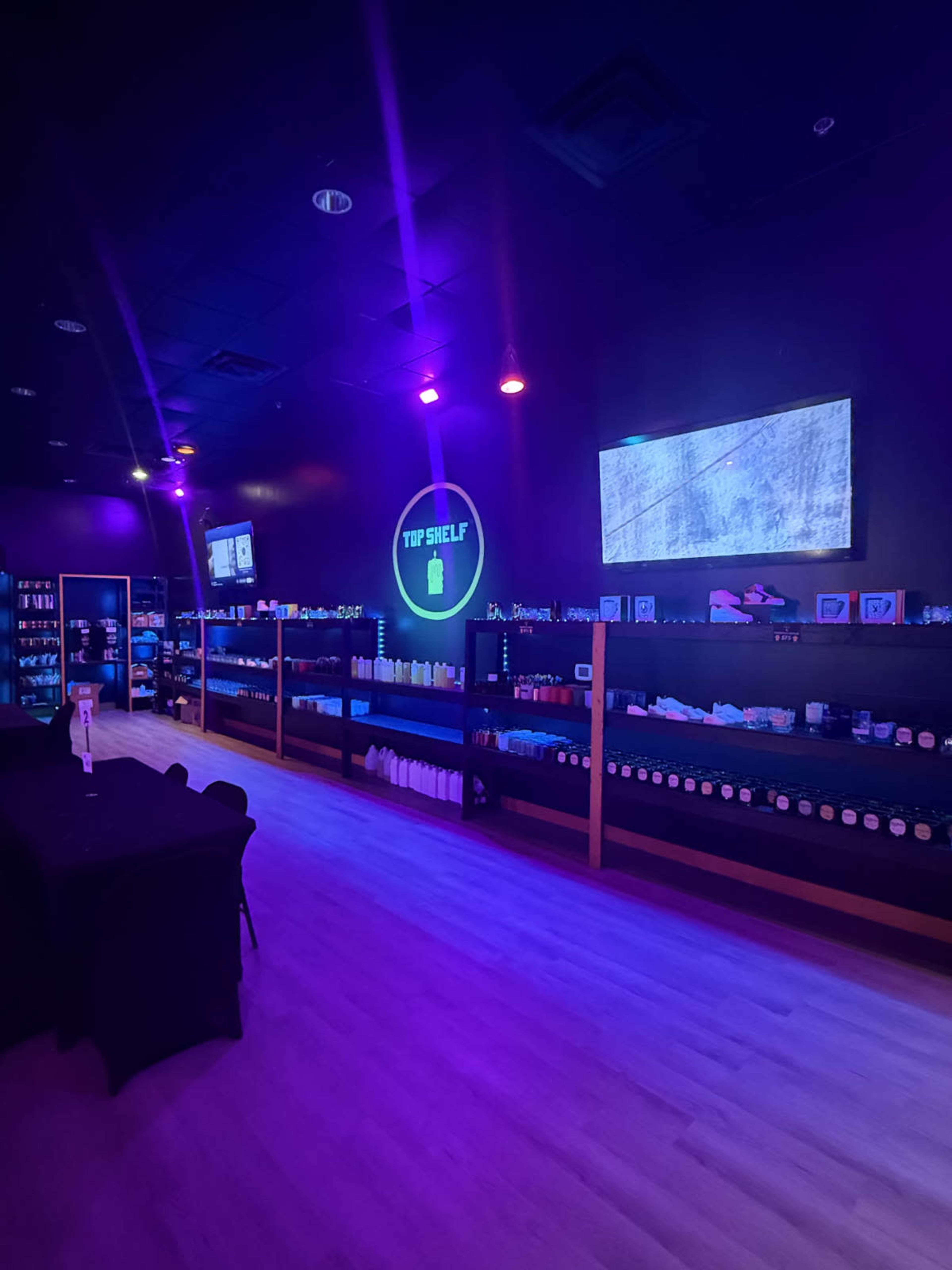 The image shows the interior of a store featuring black shelves filled with various bottles and products, illuminated by purple and white lights.