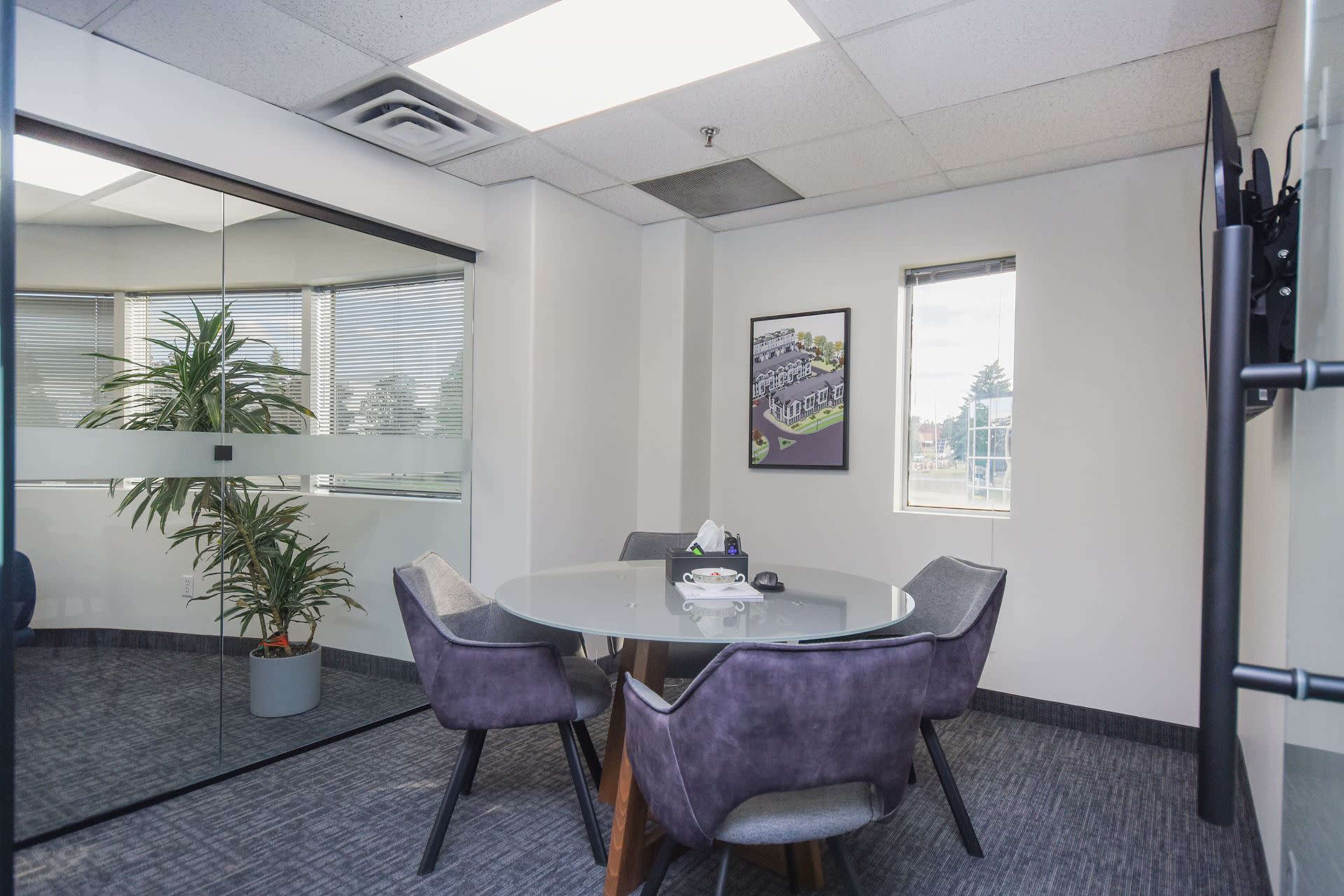 The image shows a modern office conference room with a round glass table, gray upholstered chairs, and a potted plant near a window.