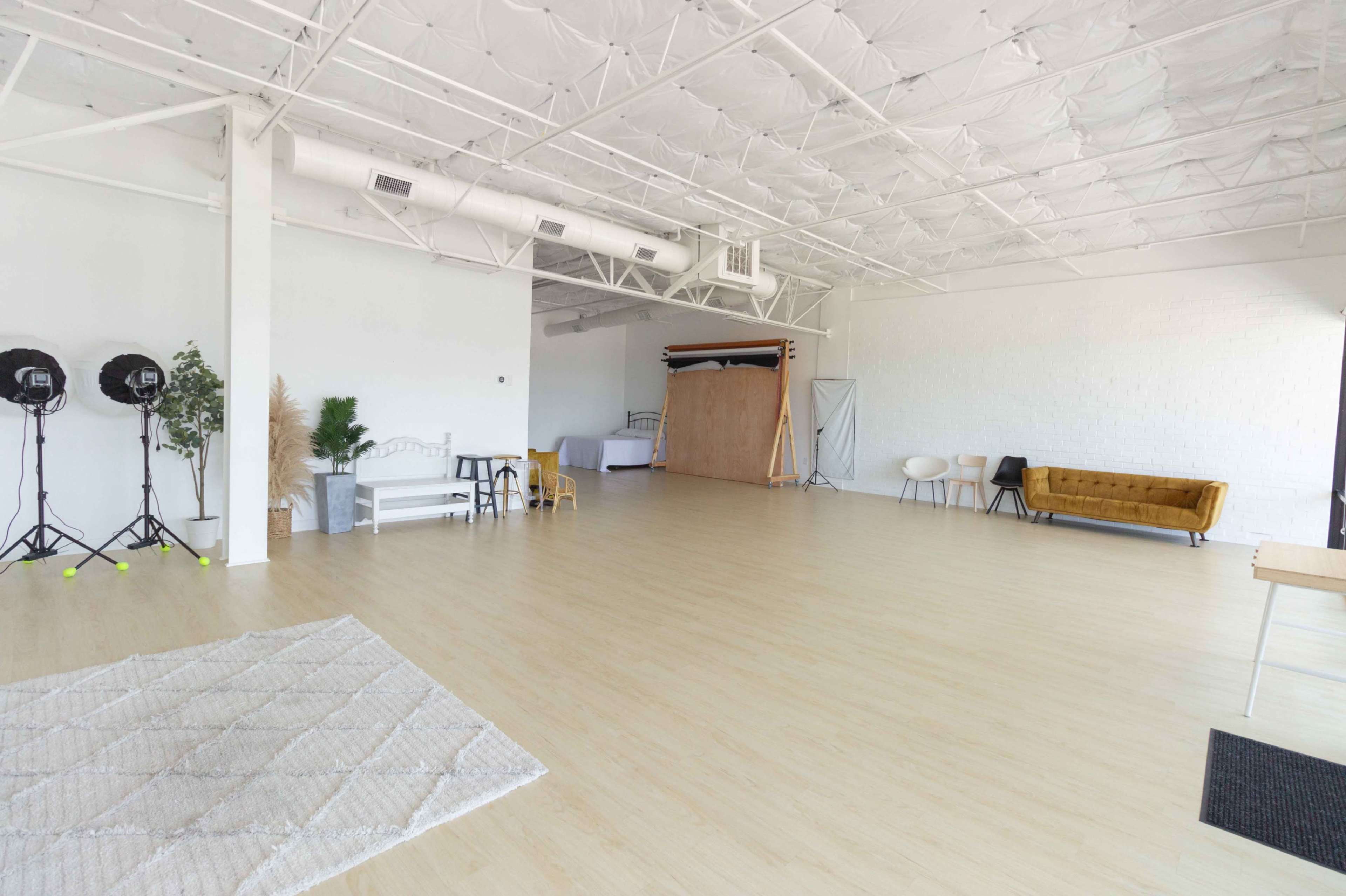 A spacious studio with wooden floors, two lighting setups, a couch, a rug, and various props along the walls.