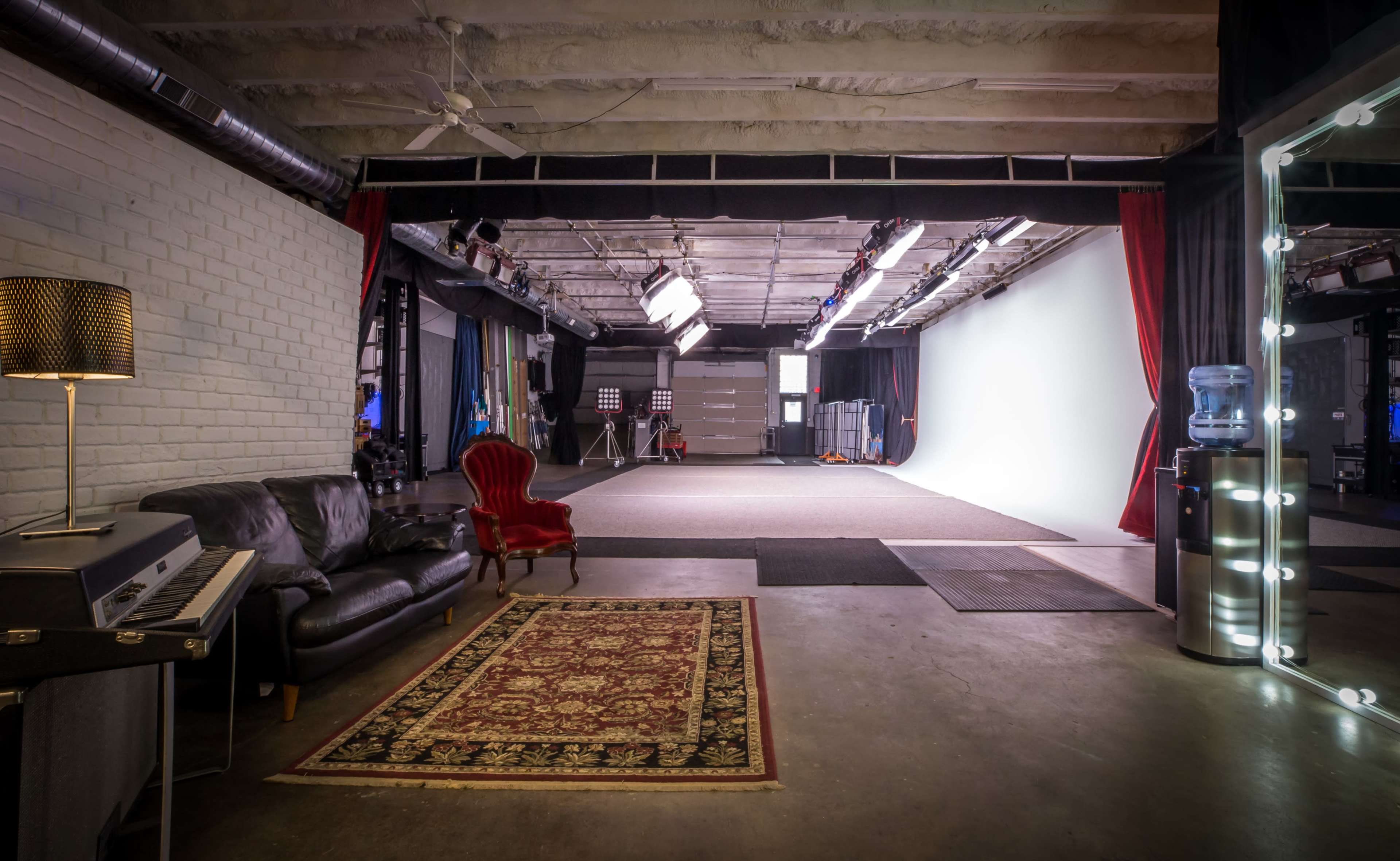 A spacious film studio with a seating area featuring a black couch and a red armchair, alongside a piano, and a large white backdrop illuminated by studio lights.