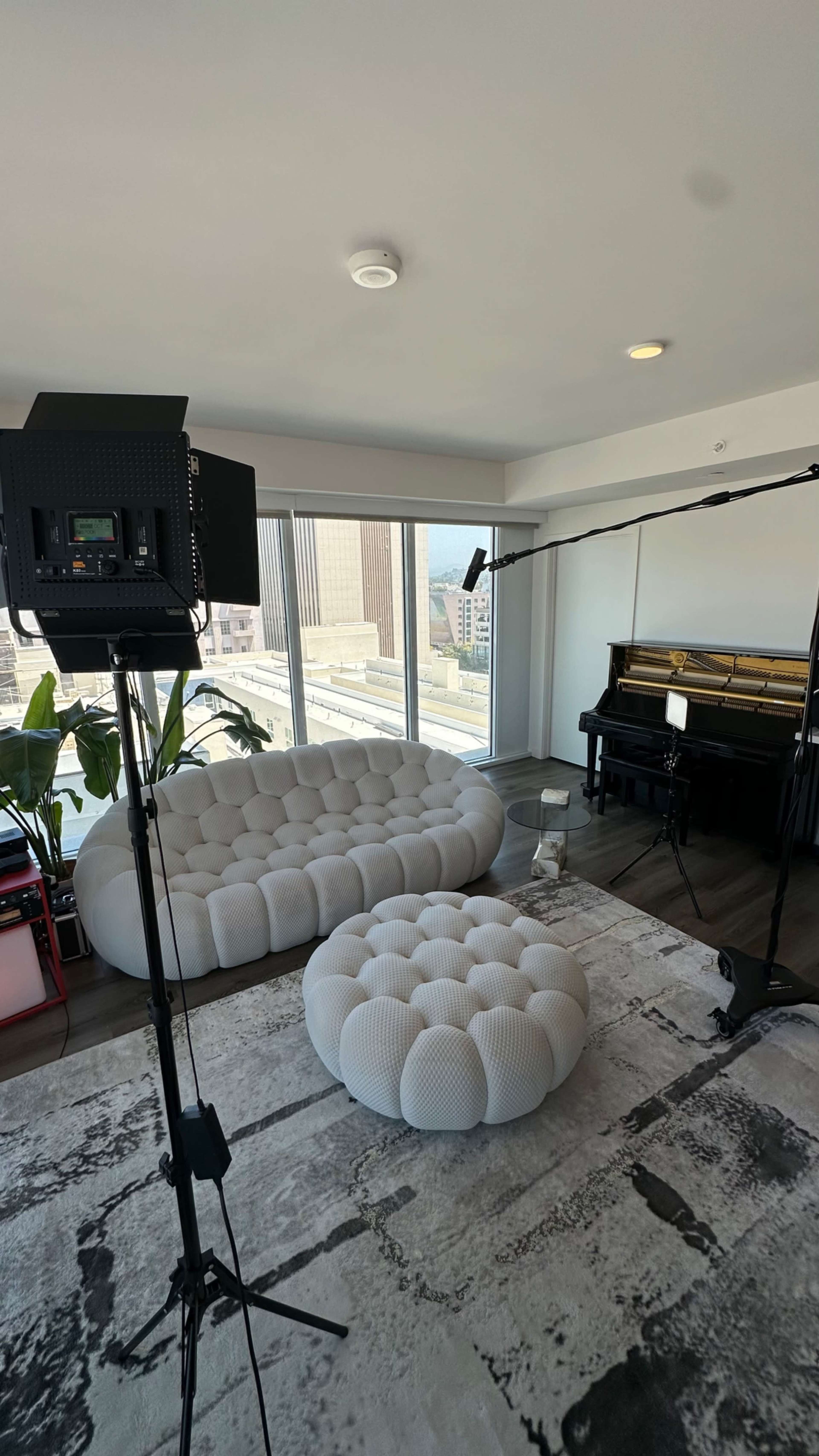 A well-lit living space features a white tufted sofa and ottoman, along with a piano and filming equipment set up near large windows overlooking the city.