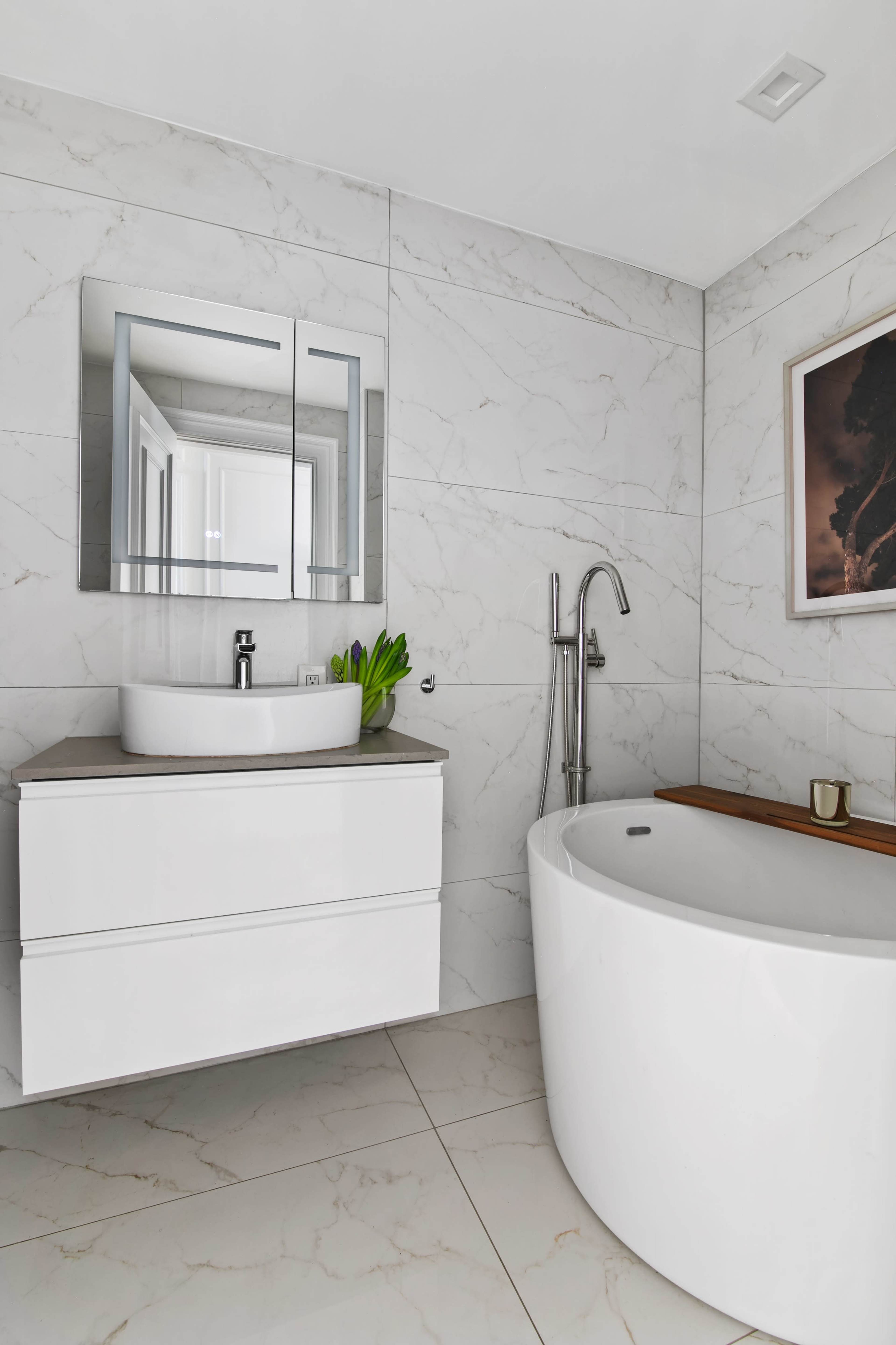 The image shows a modern bathroom featuring a white freestanding bathtub, a sleek vanity with a vessel sink, and marble-patterned tile walls.