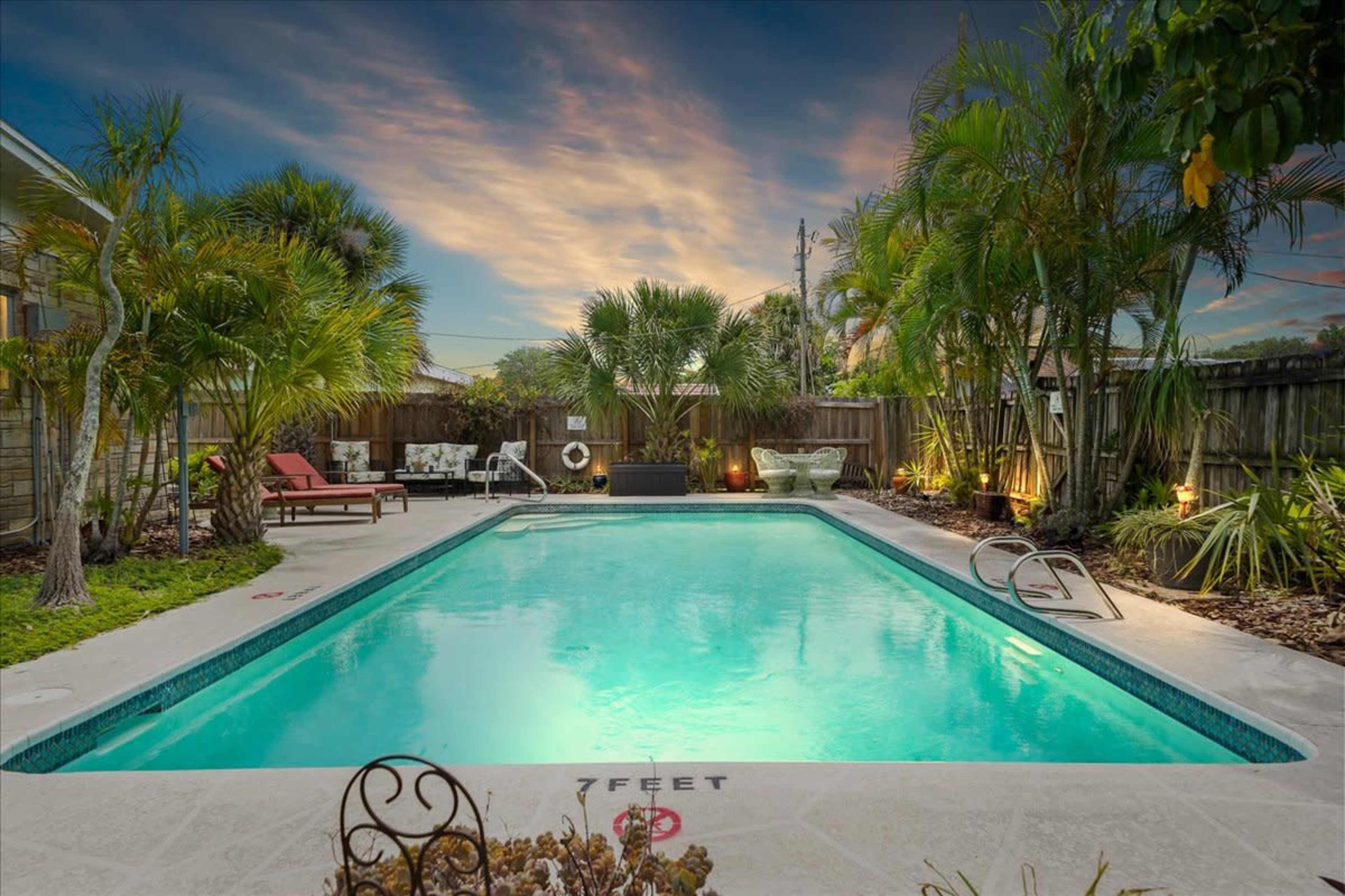 A rectangular swimming pool surrounded by palm trees and lounge chairs under a colorful sunset sky.