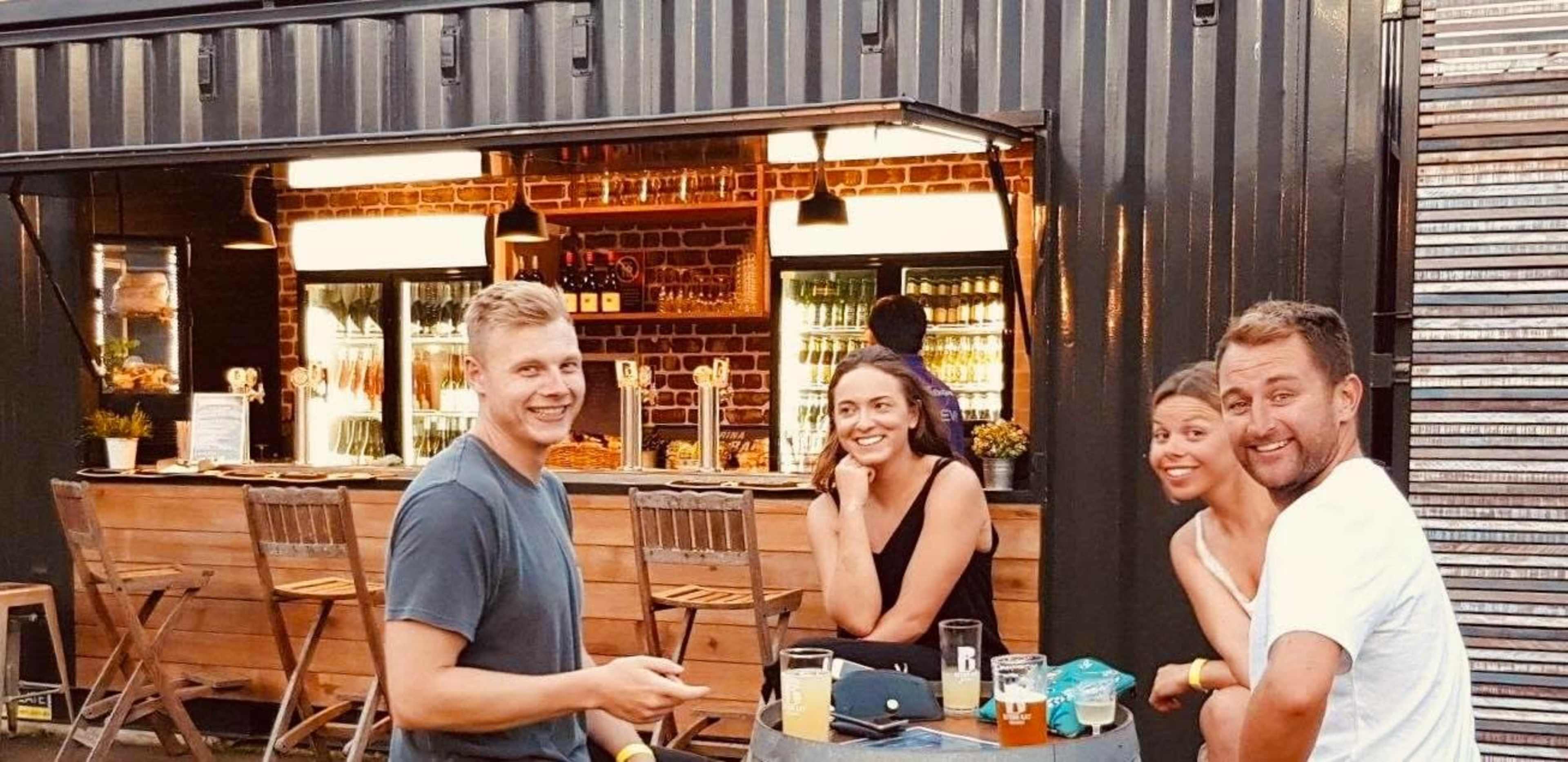 Four people are sitting at a table in front of a bar with a wooden counter and shelves stocked with drinks.