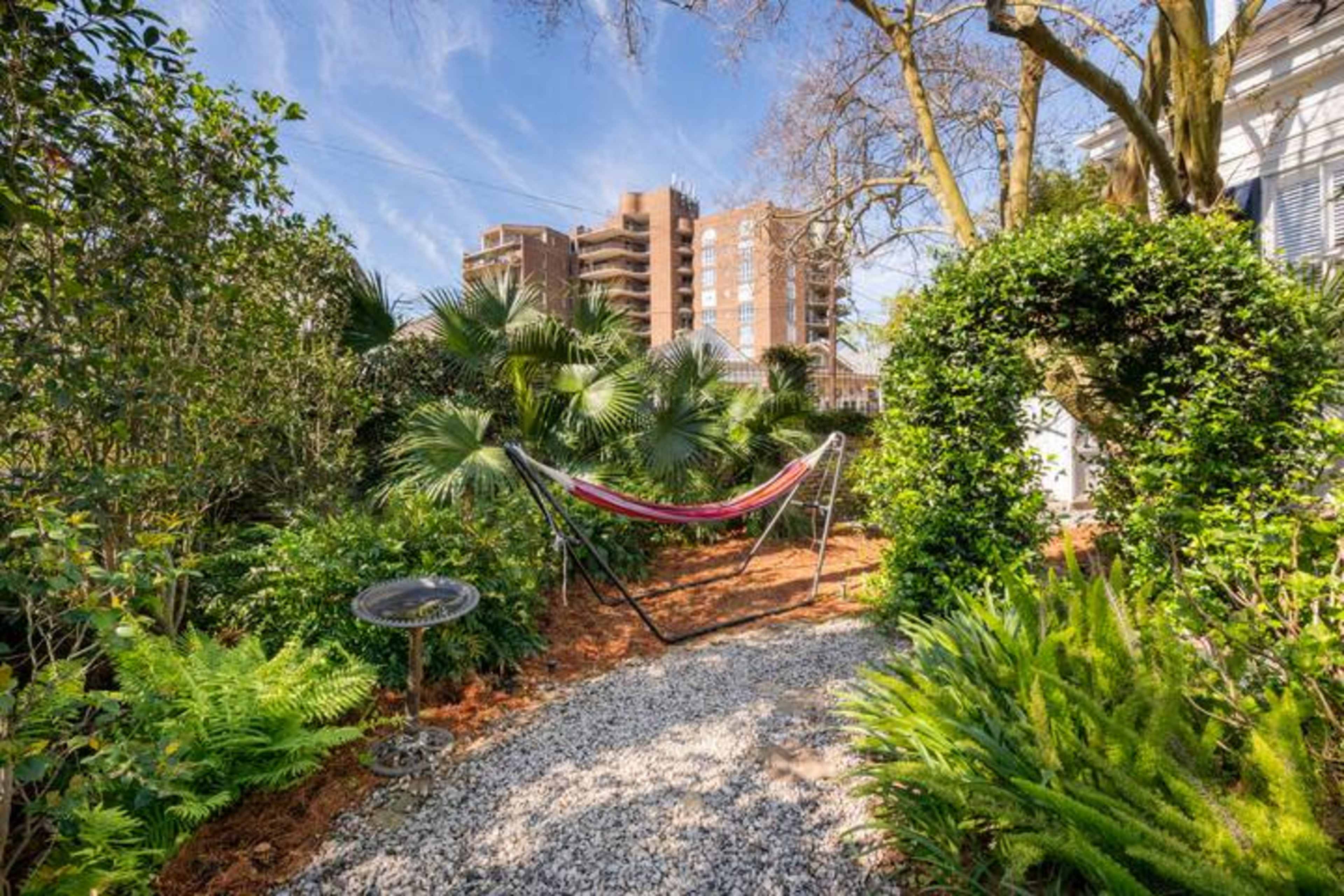 A hammock is set among lush greenery and palm plants in a garden, with tall buildings visible in the background.