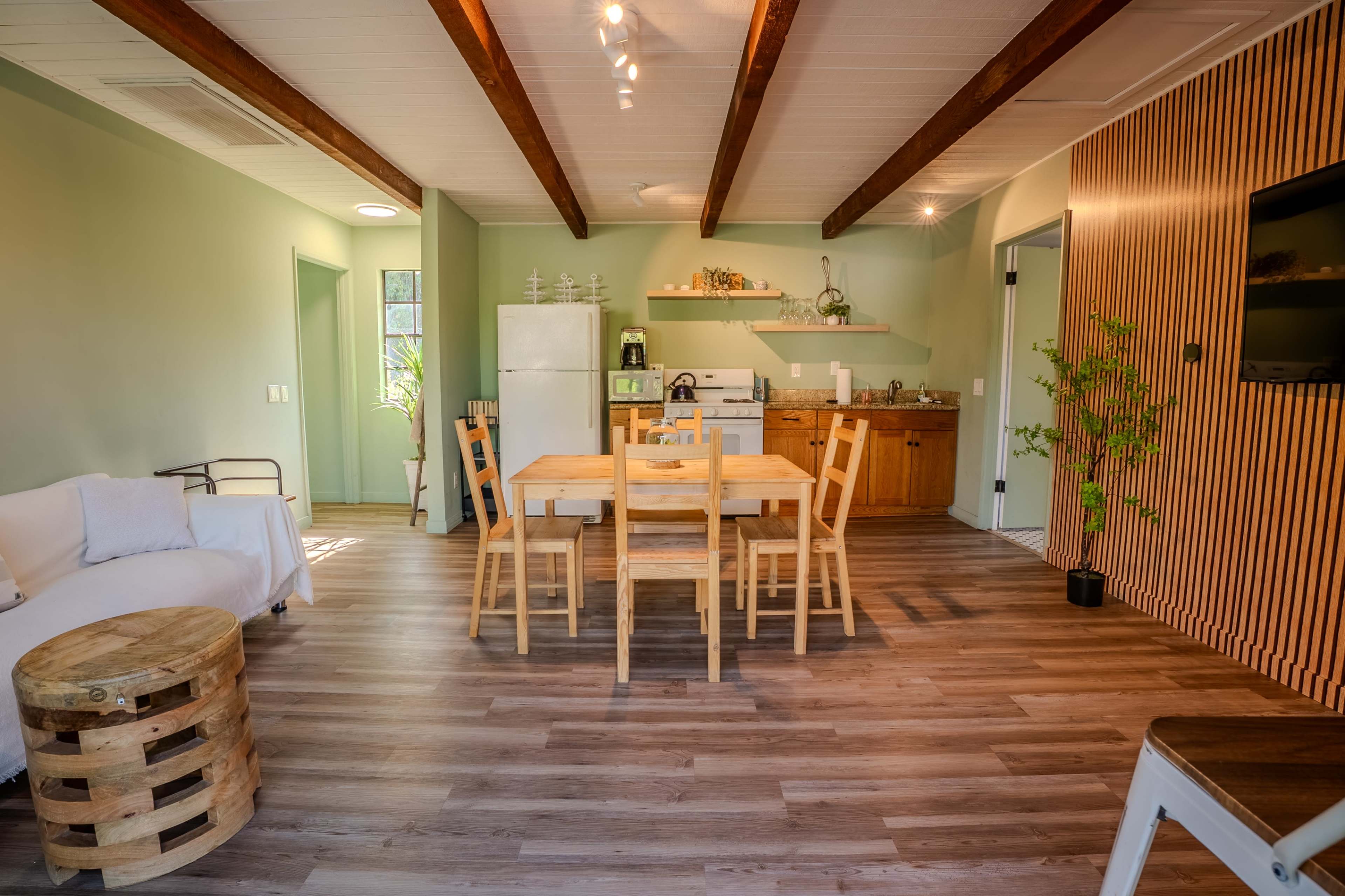 The image shows a modern, open-concept living space with a wooden dining table, chairs, kitchen area, and a small seating arrangement.