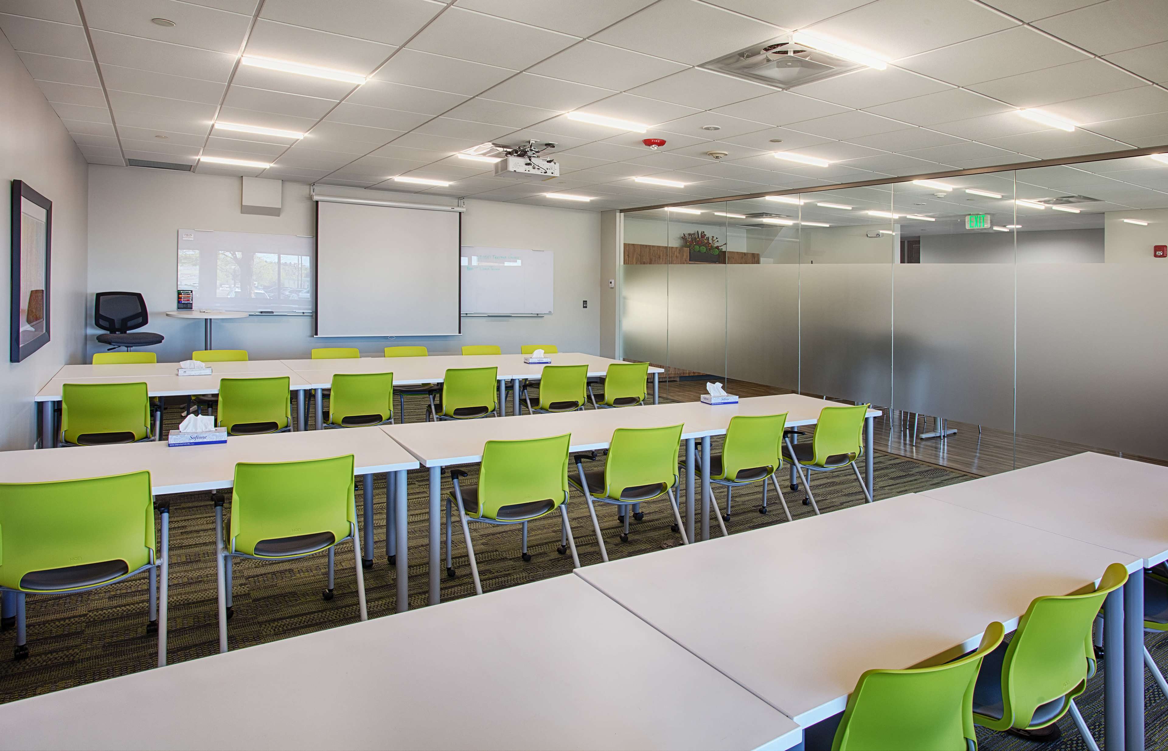 A classroom features several white tables arranged in rows with green chairs and a whiteboard at the front.