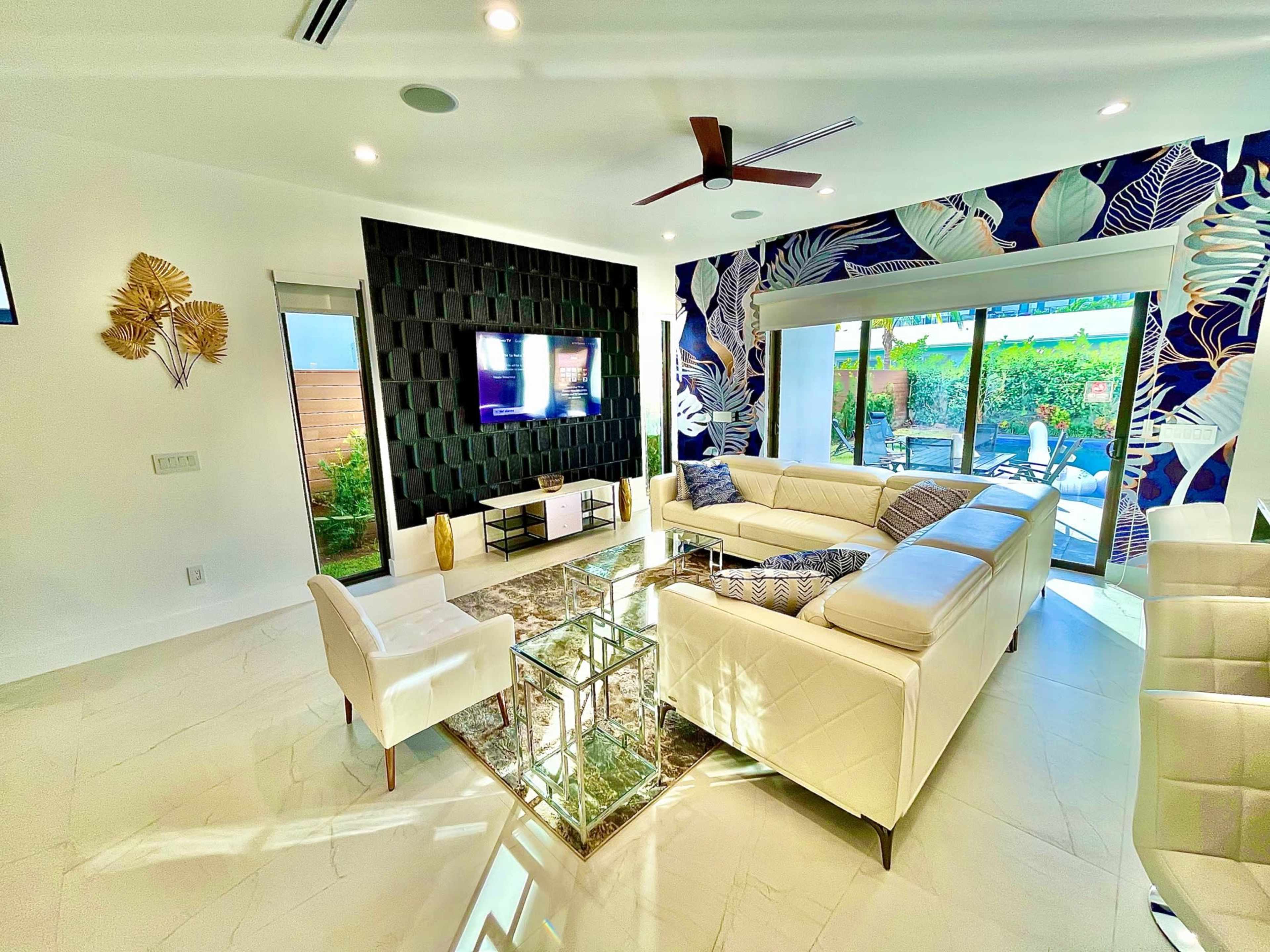 The image shows a modern living room with a white sectional sofa, a glass coffee table, and a large television mounted on a textured black wall, featuring decorative plants and a vibrant floral wallpaper in the background.