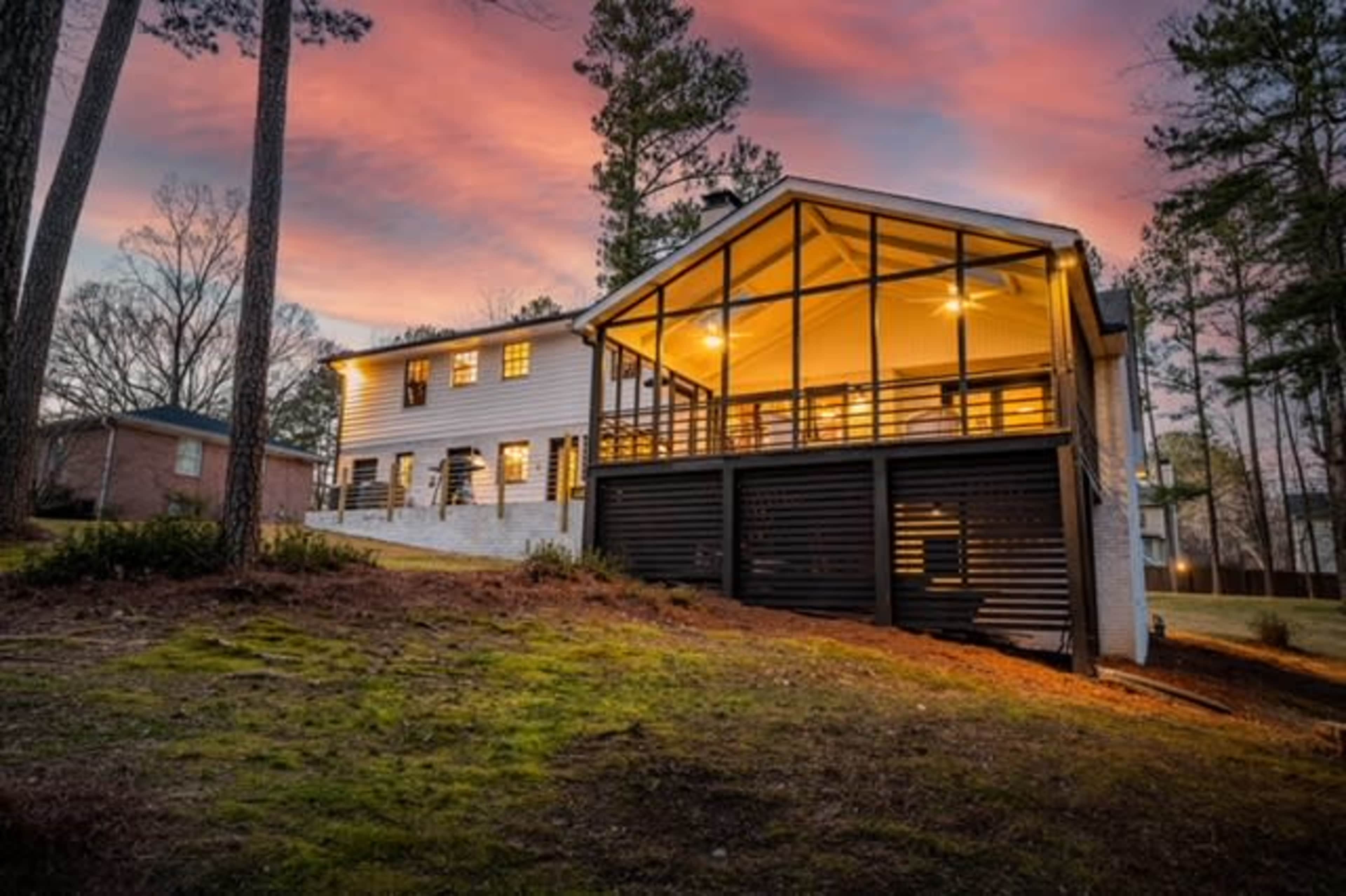A two-story house with a large deck and numerous windows is set against a colorful sunset sky.