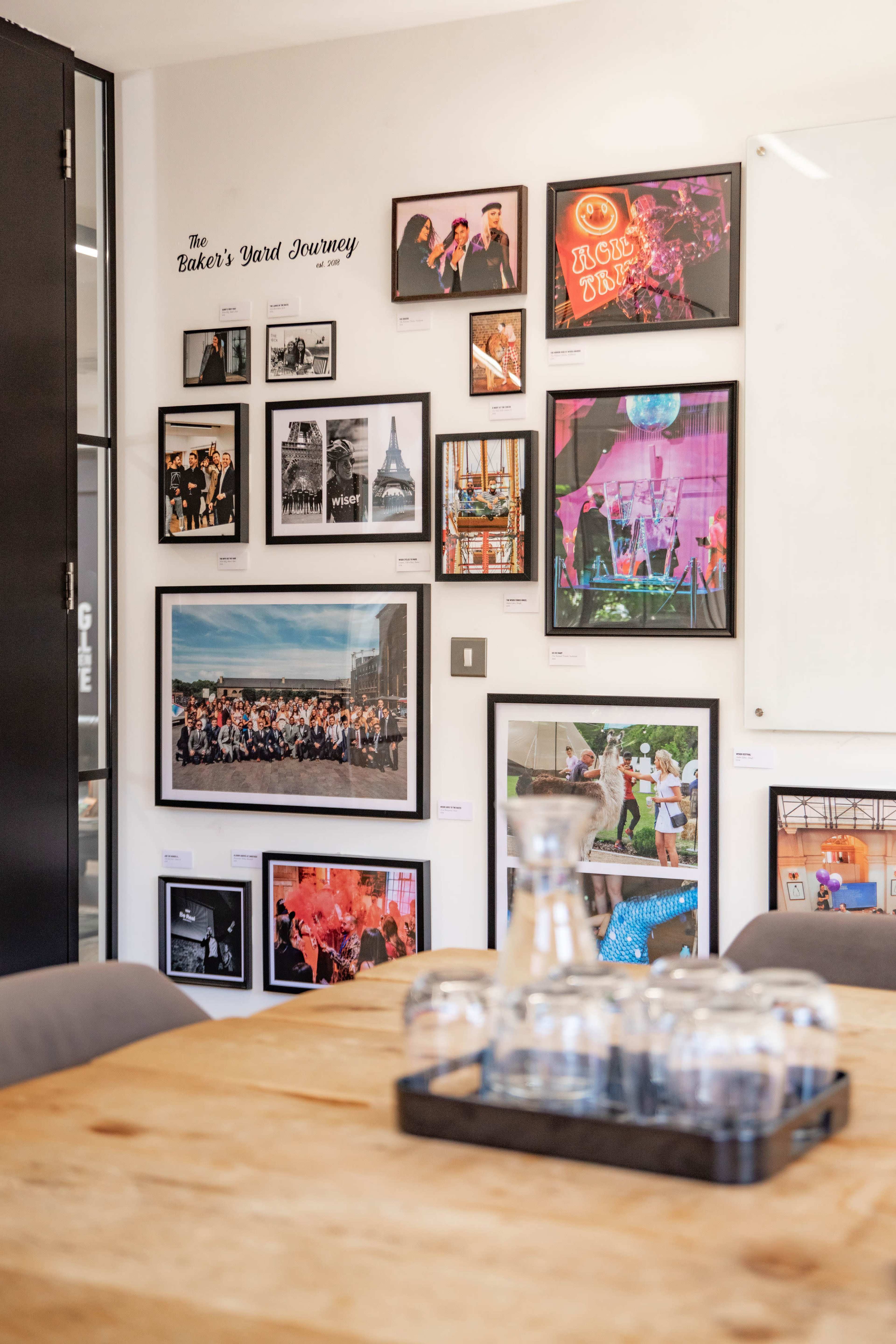 A gallery-style display of framed photographs on a wall in a brightly lit room with a wooden table in the foreground.