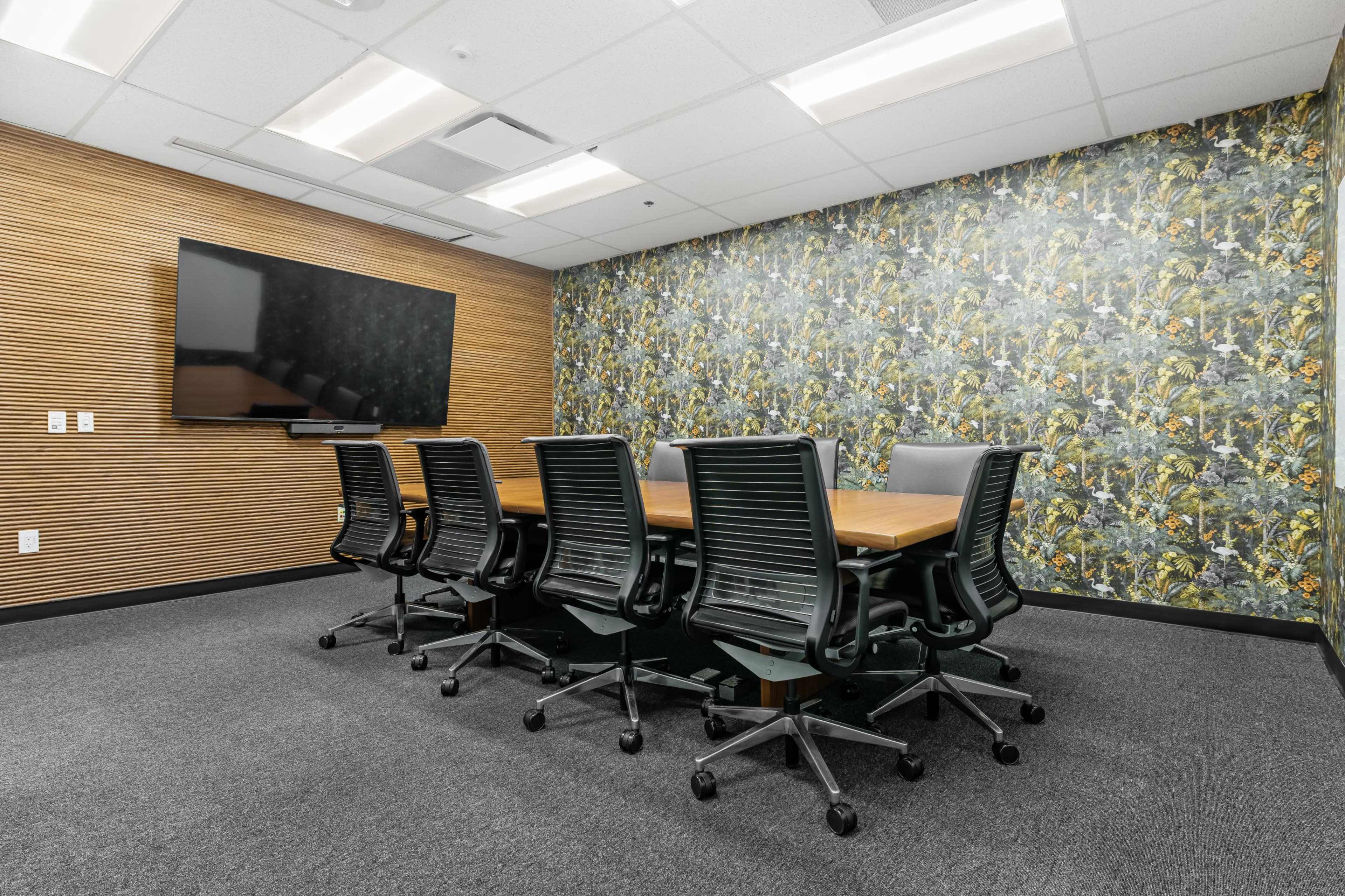 The image shows a modern conference room with a large wooden table surrounded by black swivel chairs, a flat-screen TV mounted on a wooden wall, and a floral wallpaper accent.