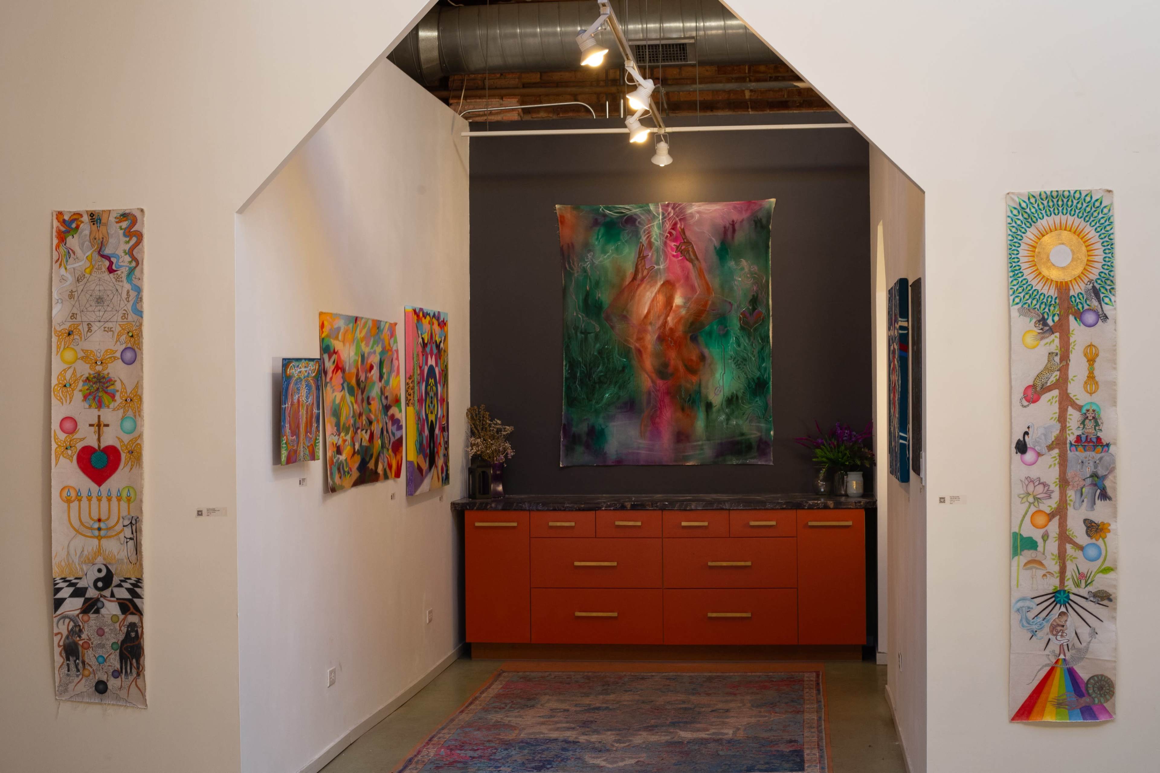 The image shows an art gallery with colorful paintings on the walls, featuring an orange cabinet and a floral arrangement in the center.