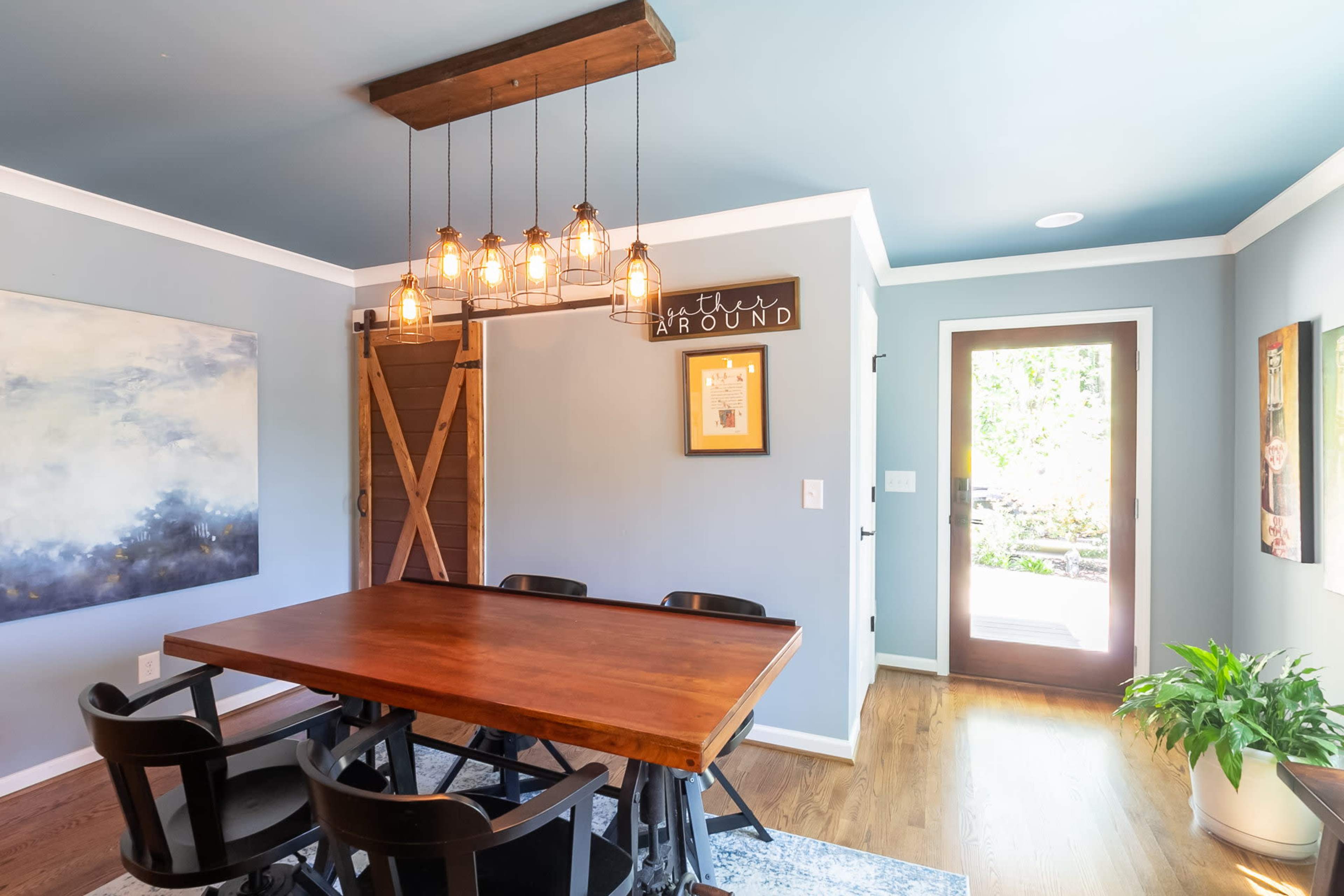 A dining area featuring a wooden table surrounded by black chairs, a light fixture with multiple bulbs overhead, and a sliding wooden door leading to an outdoor space.
