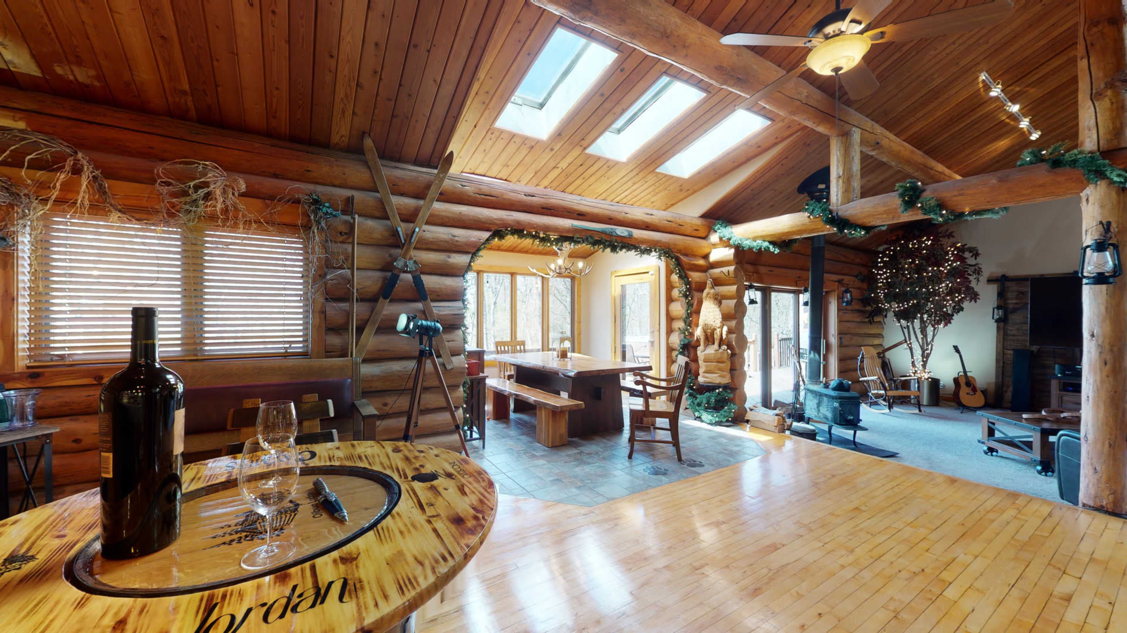 A spacious log cabin interior features a dining area with a wooden table and chairs, large windows, and a living space with musical instruments and holiday decorations.