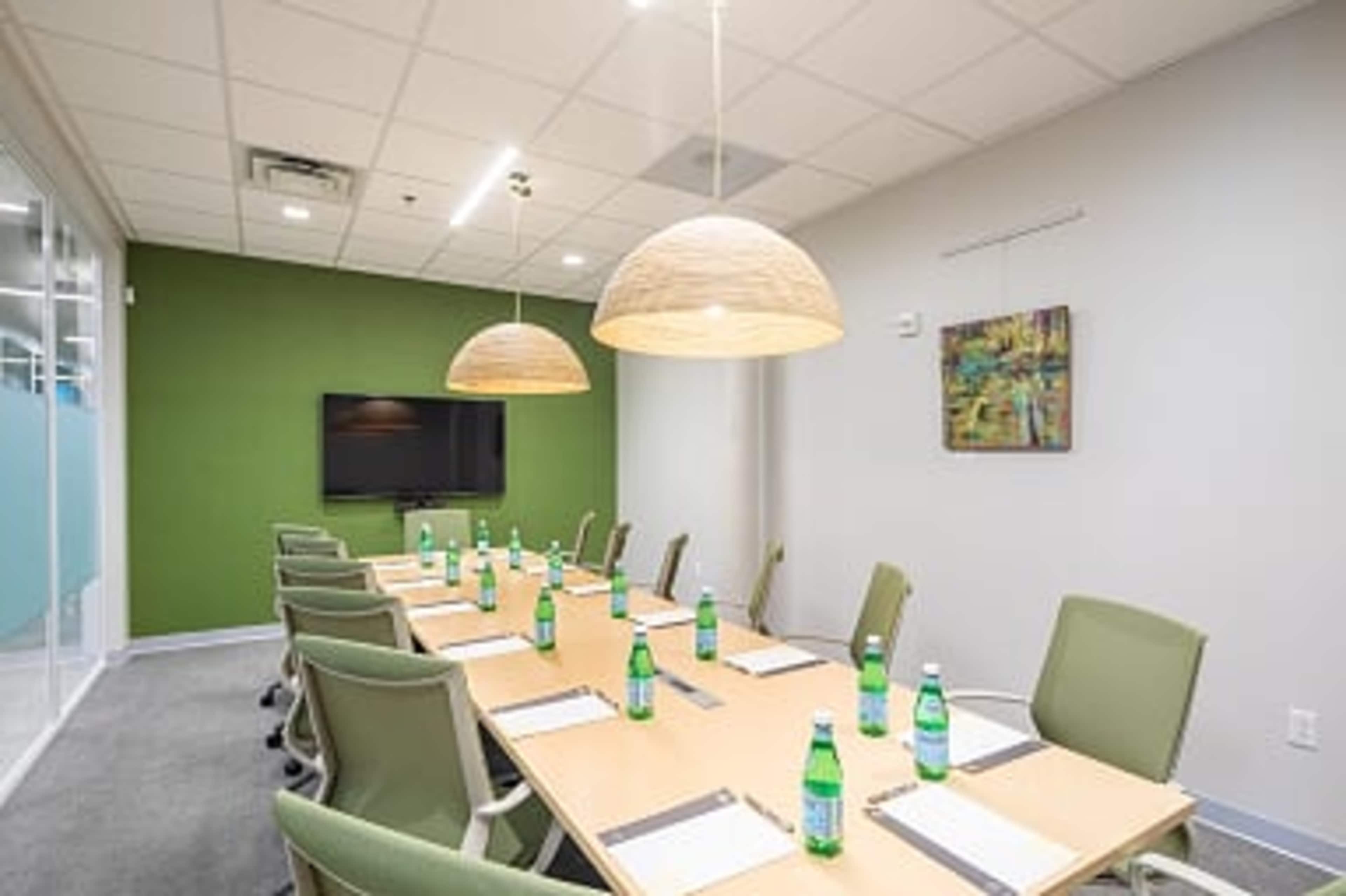 A modern conference room features a long table set with water bottles, surrounded by green chairs and illuminated by pendant lights.