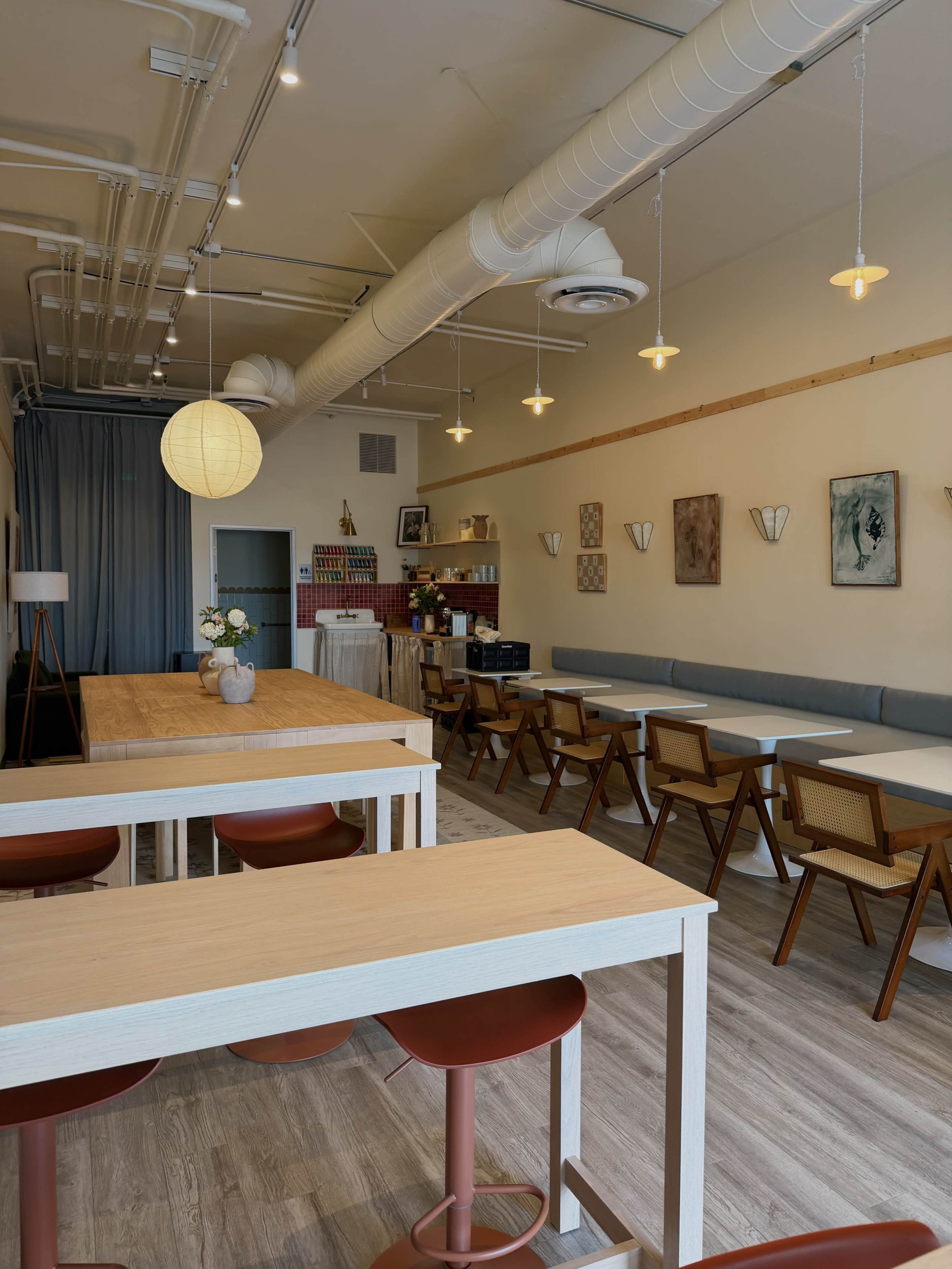 The image shows a minimalist café interior with wooden tables, chairs, and soft lighting from pendant bulbs.