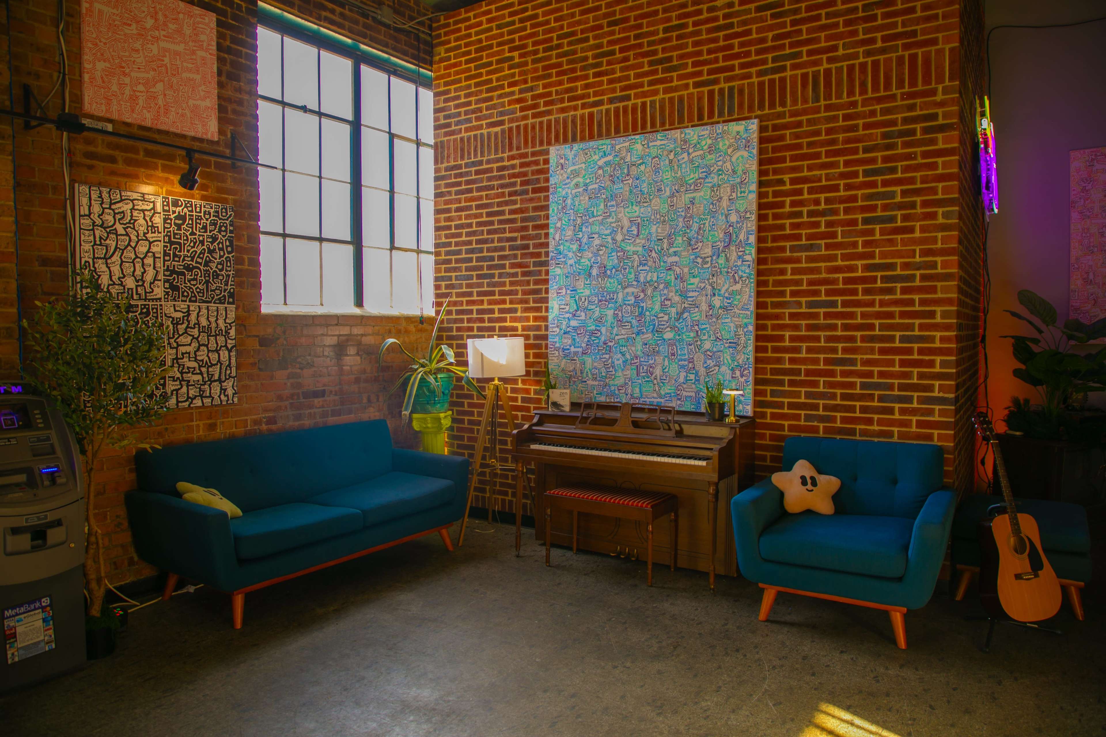 The image features a cozy seating area with two teal sofas, a piano, a colorful wall art piece, and various plants against a brick wall, with a large window allowing natural light.